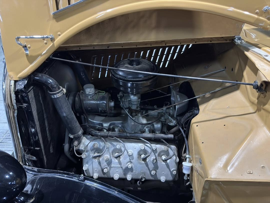 Open hood of a mustard-yellow vintage car showing the exposed inline engine, carburetor, radiator, and wiring details in the engine bay