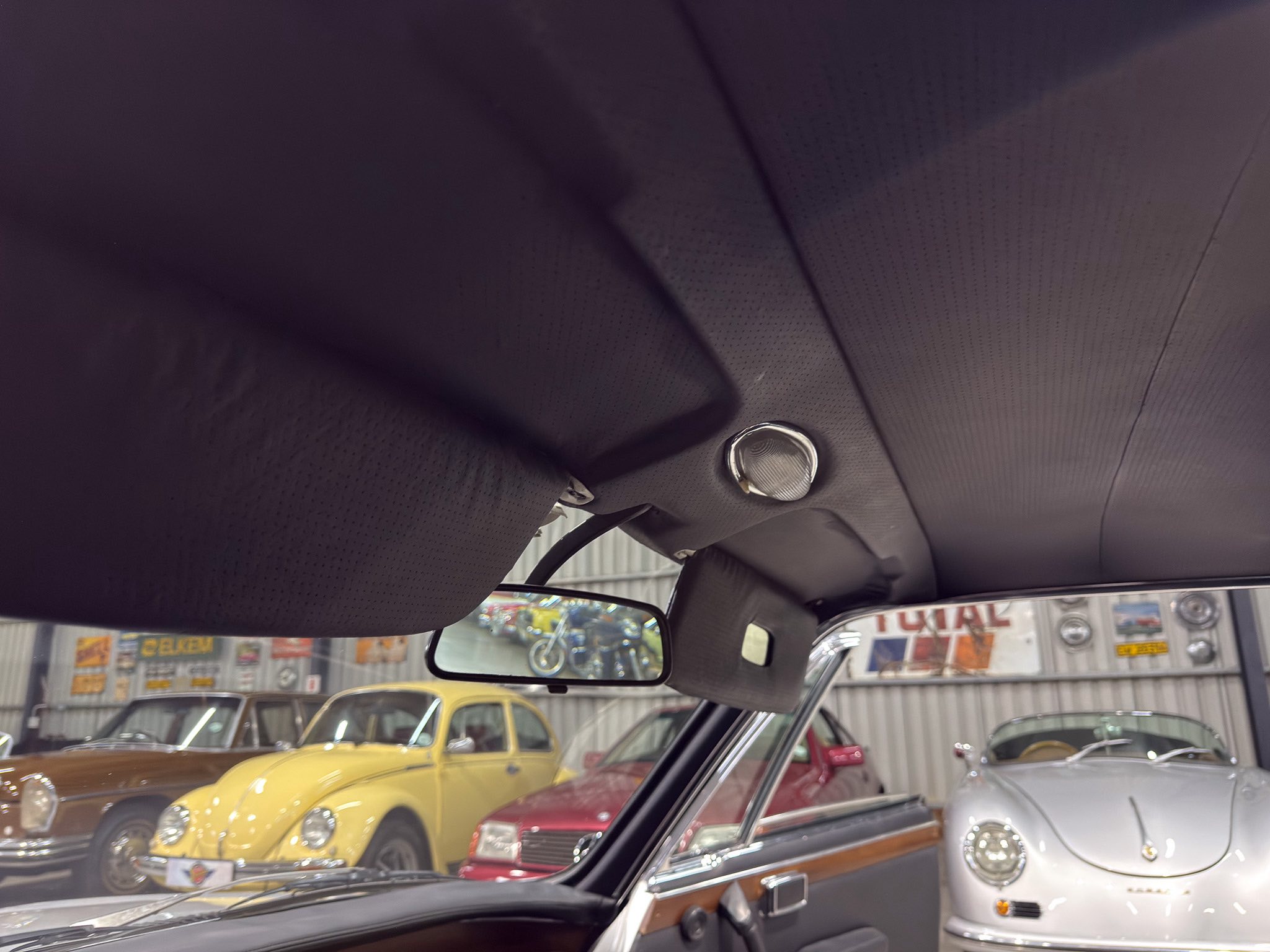 View from inside a classic car looking toward a showroom full of vintage cars, with a dark padded headliner and rearview mirror in the foreground.
