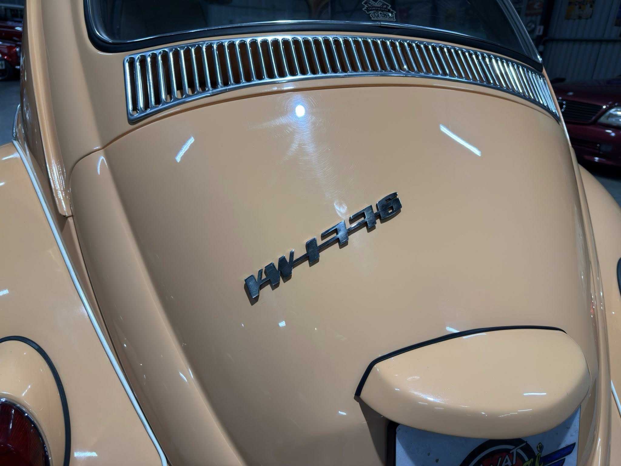 Rear section of a beige vintage car showing a curved hood, chrome grille vents, and partial badge lettering along the front edge of the trunk/hood area.