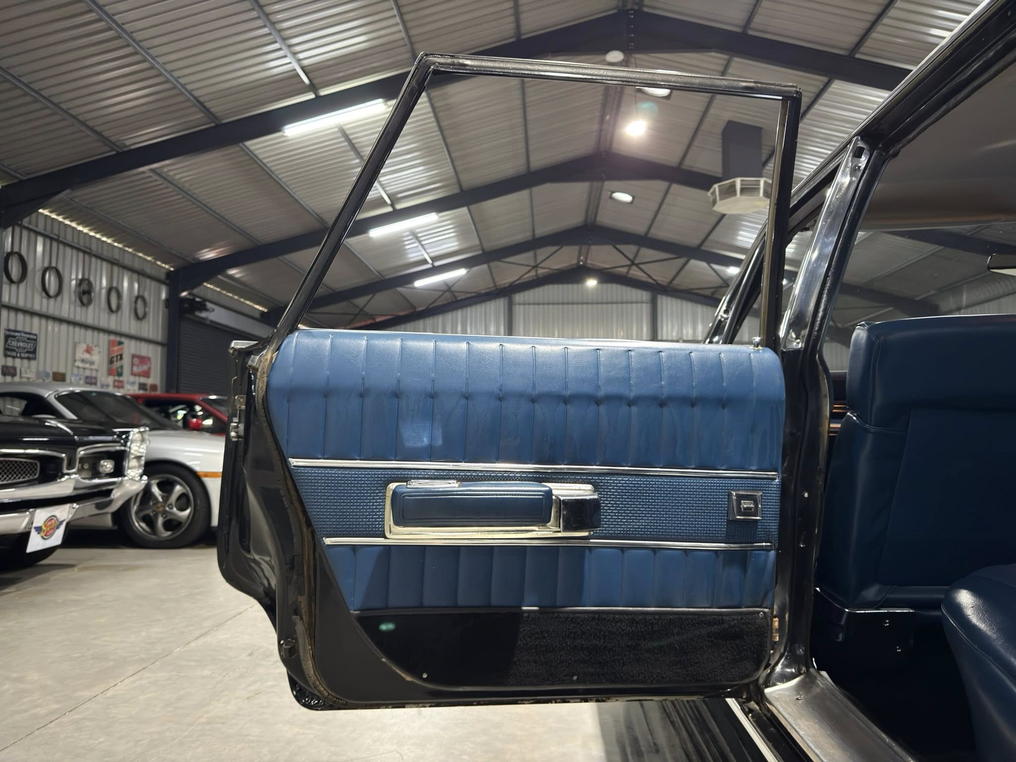 Open door of a blue vintage car showing the tufted blue interior panel inside a showroom or workshop with other cars in the background.