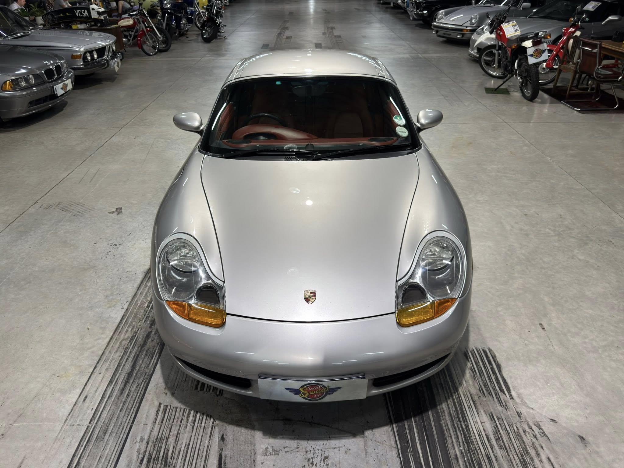 Front view of a silver Porsche in a showroom, with motorcycles and other cars lining the space.