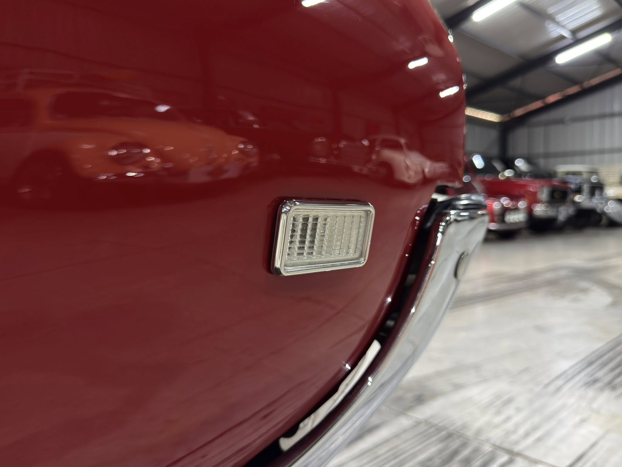 Close-up of a red car's rectangular taillight on the rear corner, with a blurred garage and other vehicles in the background.