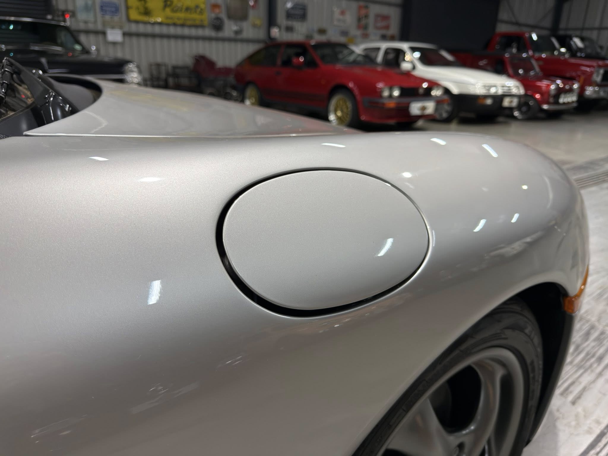 Close-up of a silver car's fuel door on the side, with a showroom full of red and white cars in the blurred background.