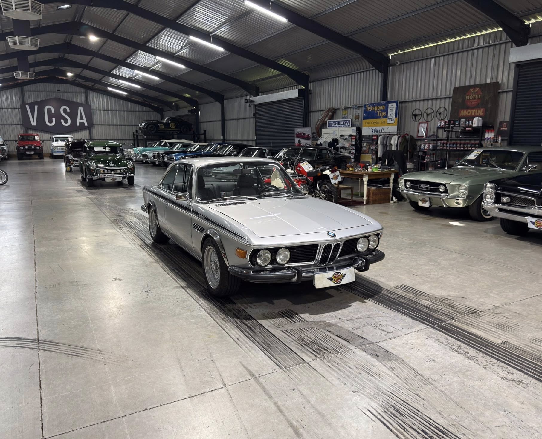 Indoor showroom filled with vintage cars; silver BMW in the foreground and a large VCSA sign on the back wall