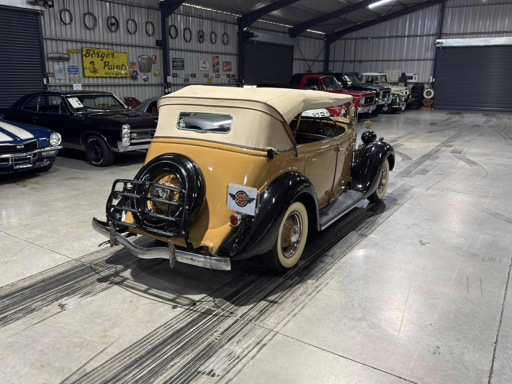 Indoor showroom of classic cars, with a tan convertible in the foreground and more vintage vehicles lined up along the wall behind it.