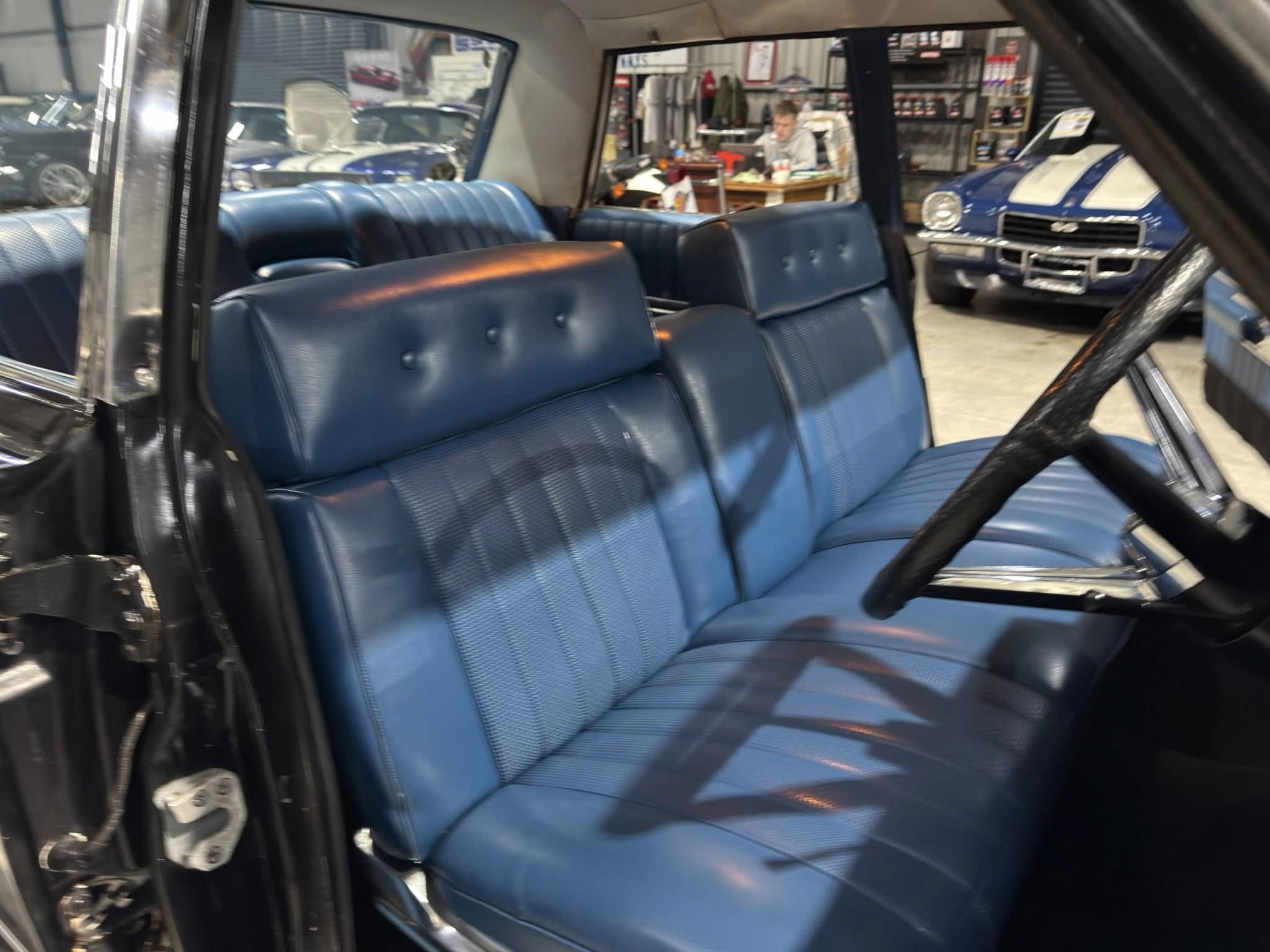 Inside a blue leather bench-seat car shown from the driver's side, with a showroom of classic cars and a person working at a desk in the background.