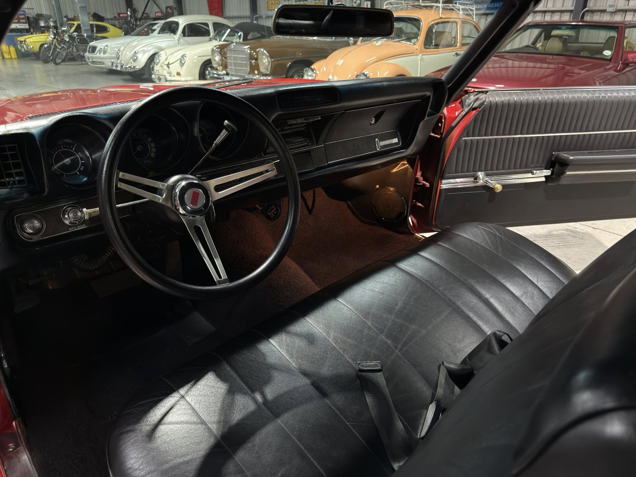Interior of a classic car showing black dashboard and steering wheel, with a row of vintage cars visible through the windshield in a showroom.