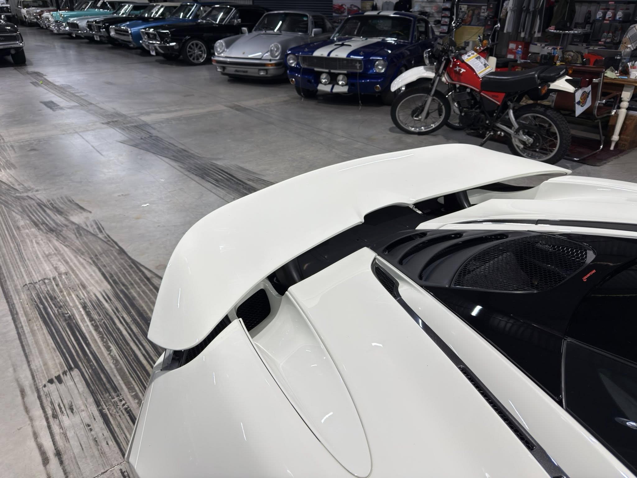 View inside a car showroom with vintage and modern vehicles lined up along the wall; foreground shows the hood of a white sports car.
