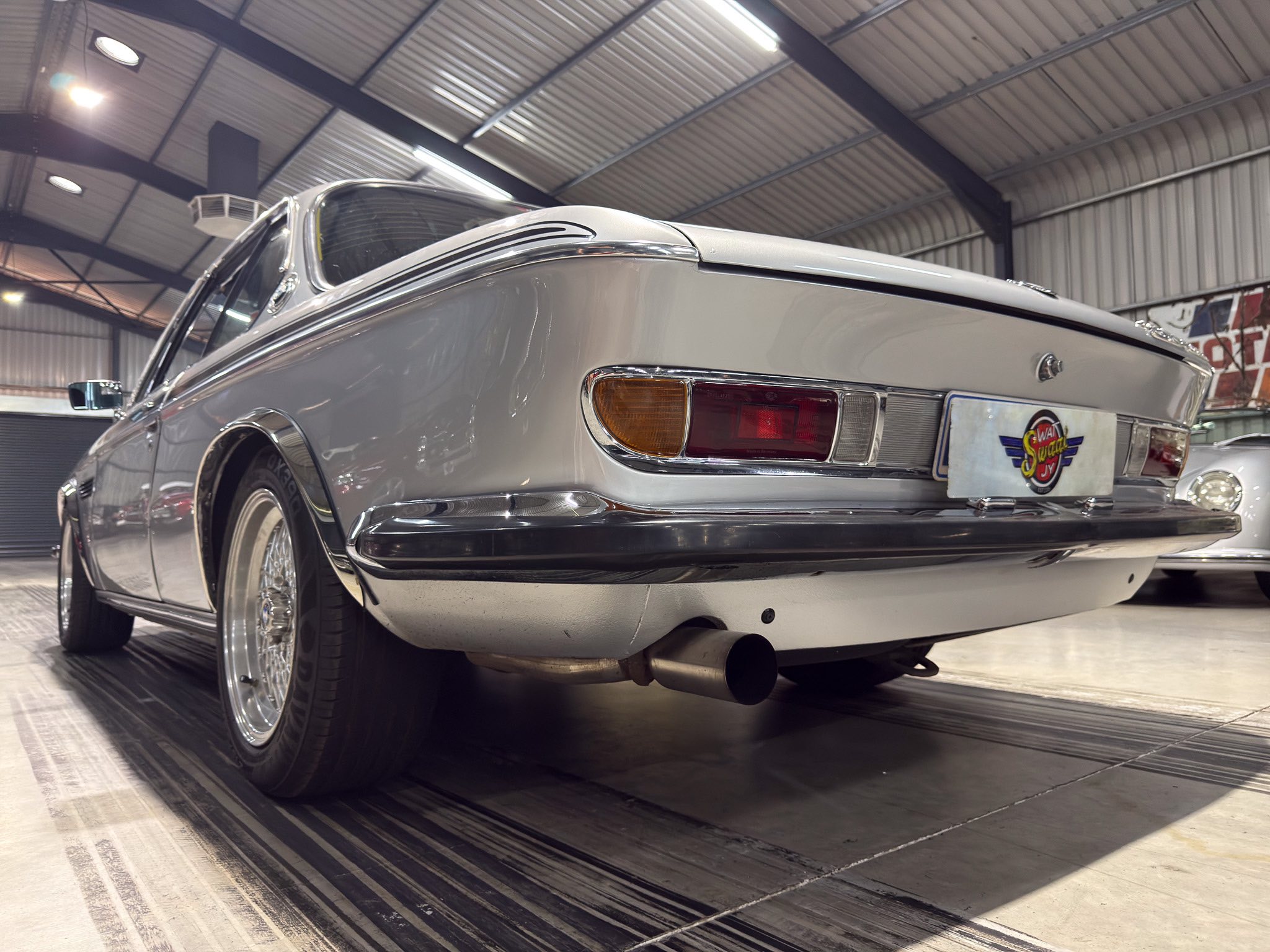 Rear three-quarter view of a vintage silver car on display in a well-lit showroom, chrome bumper visible behind the taillights.