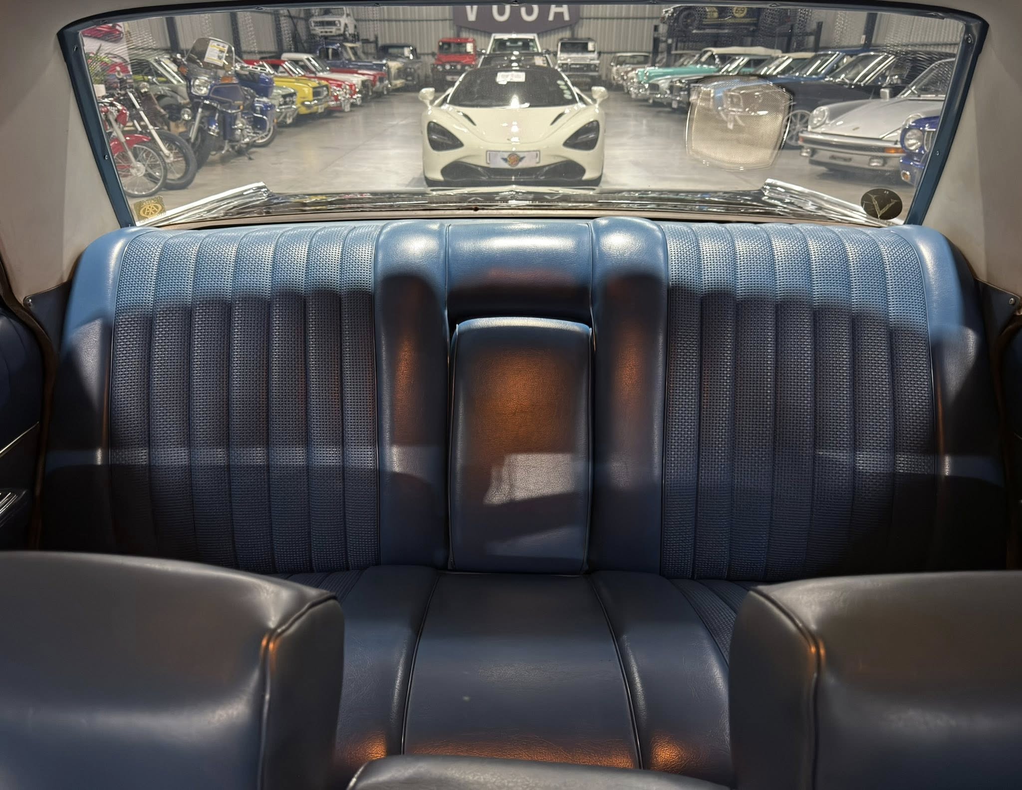 Backseat view inside a blue vintage car, showroom filled with colorful classic cars seen through the rear window center stage white sports car.