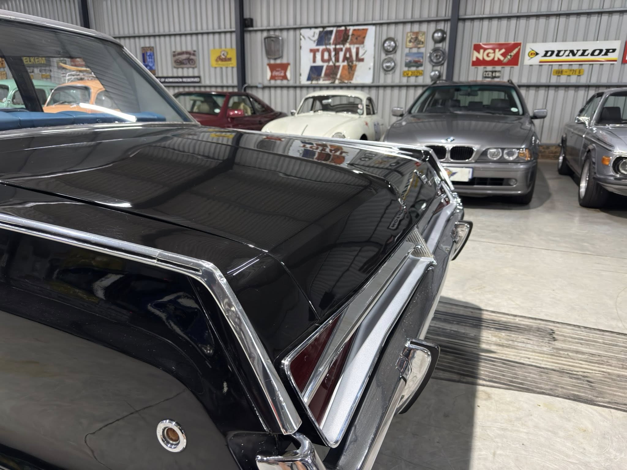 Close-up of a glossy black vintage car trunk with chrome trim in a showroom, with other classic cars and retro signs in the background.