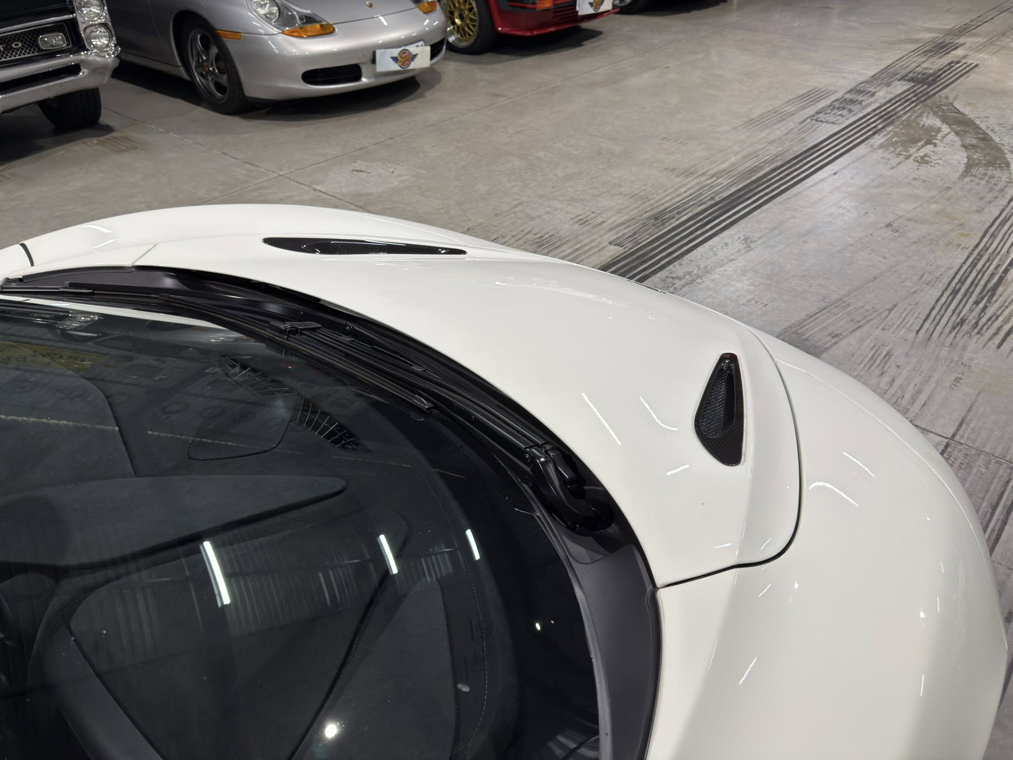 White car hood and windshield close-up in a showroom with other cars in the background.