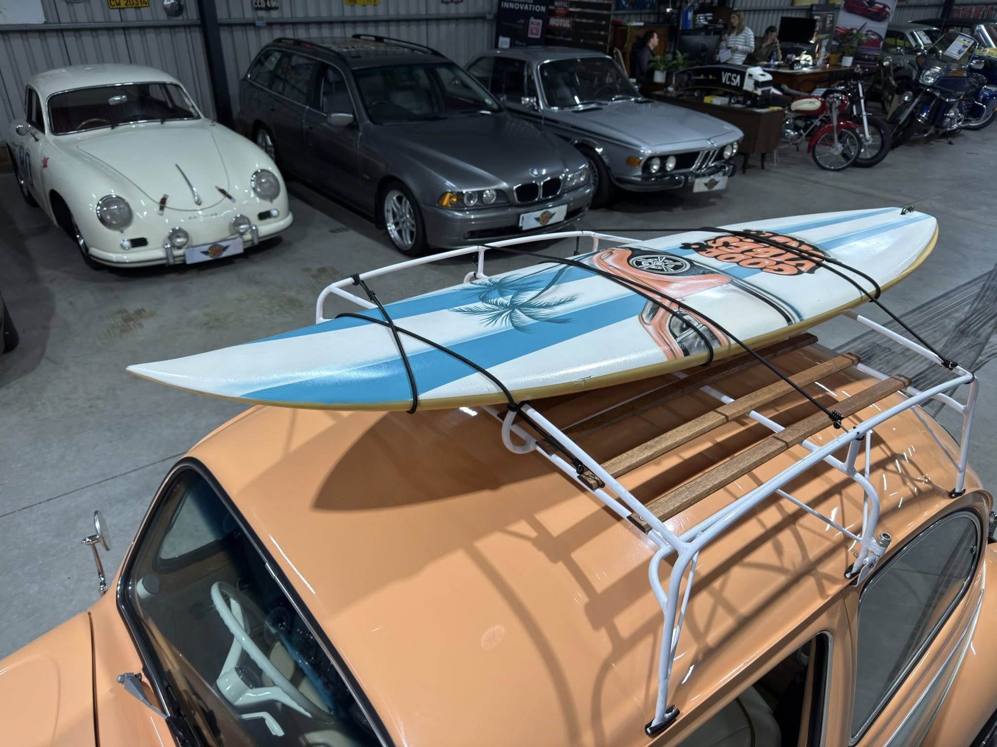 Indoor automotive showroom with a row of classic cars, incl. a white vintage Porsche, a gray sedan, and a silver BMW, plus a surfboard strapped to an orange vehicle in the foreground.
