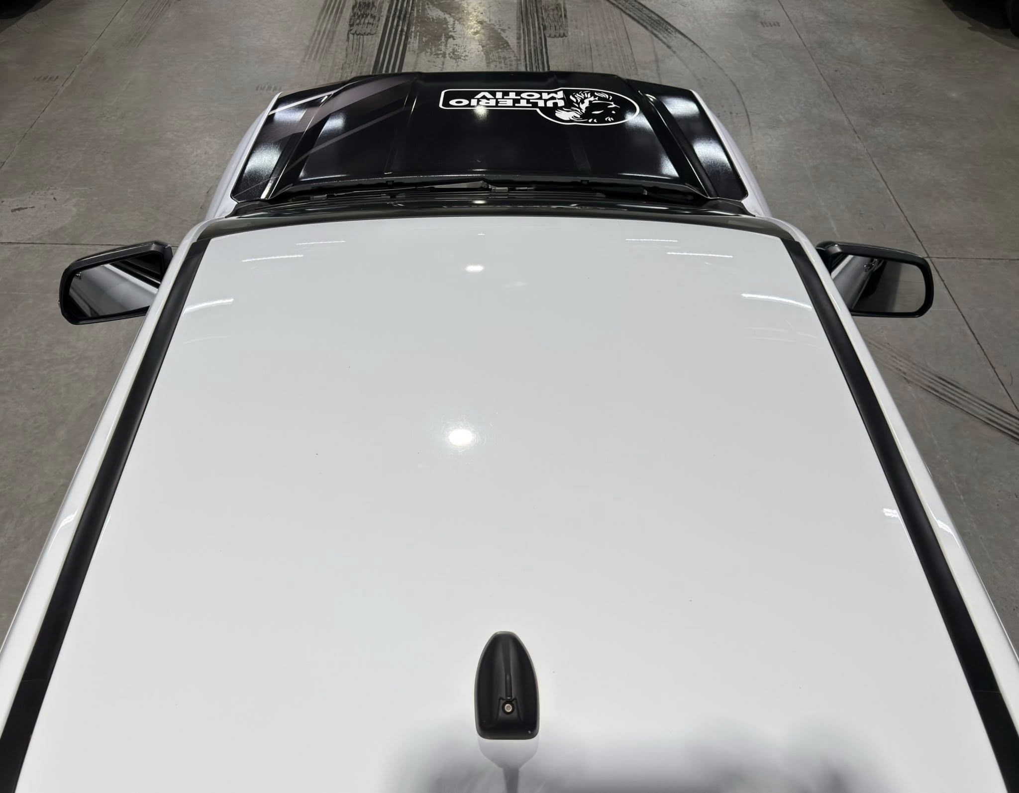 Bird's-eye view of a white car with a black panoramic sunroof and a white logo sticker centered on the glass, seen from above in a showroom.