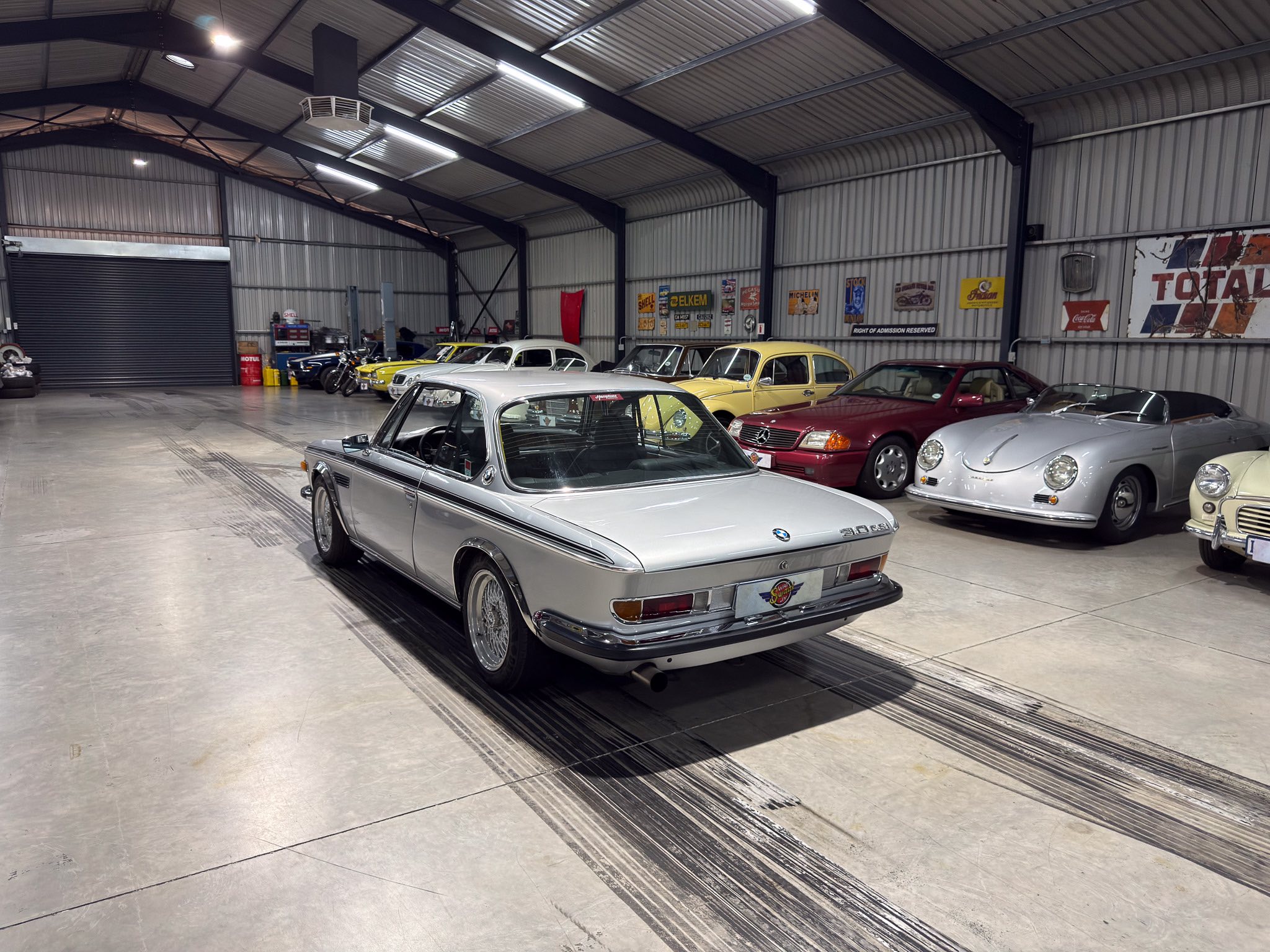 Indoor classic car showroom with a row of vintage Mercedes and Porsche convertibles inside a metal-walled warehouse.