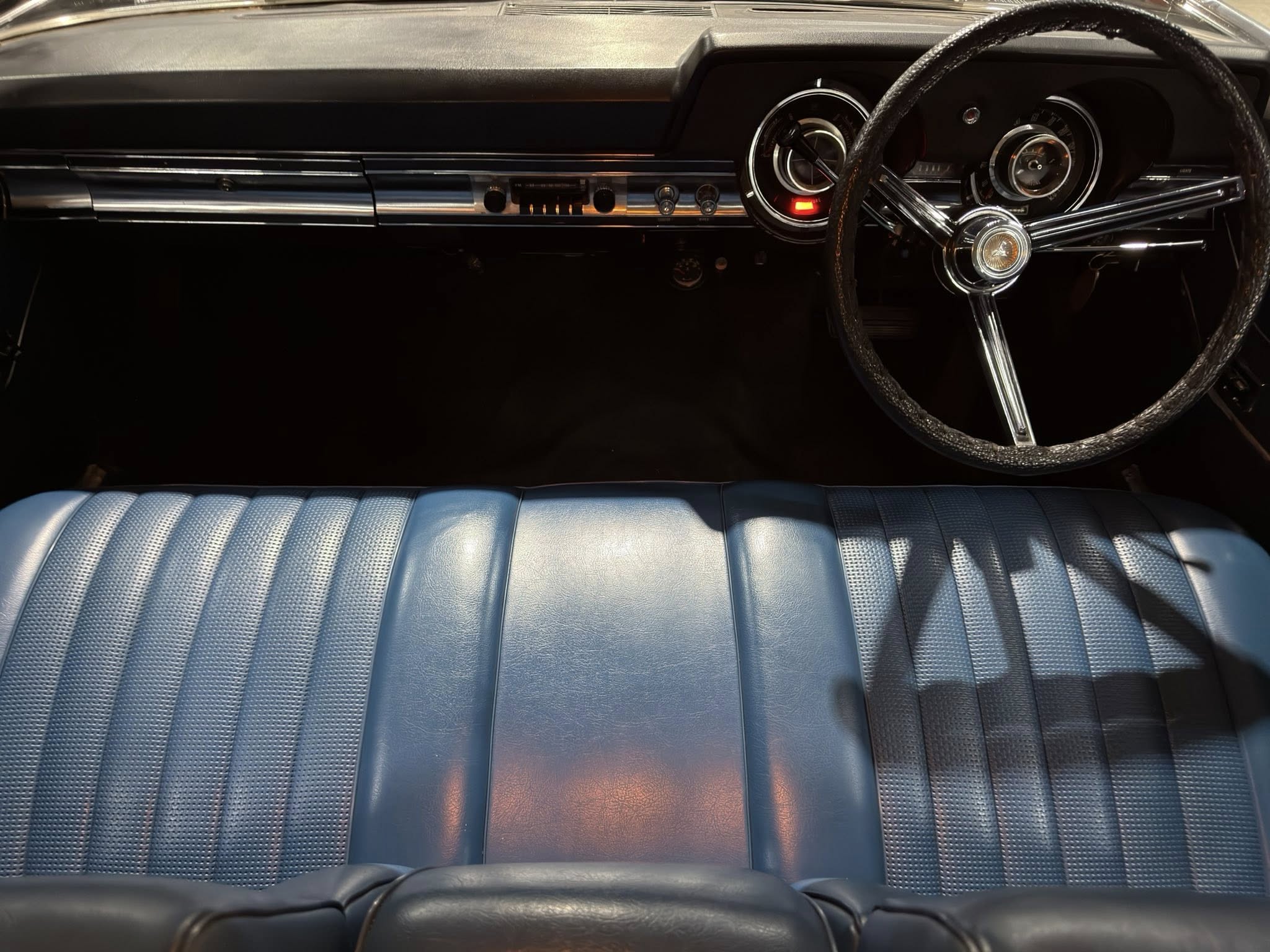 Back-seat view of a vintage car interior showing a chrome steering wheel, circular gauge cluster, and a blue bench seat in the foreground.