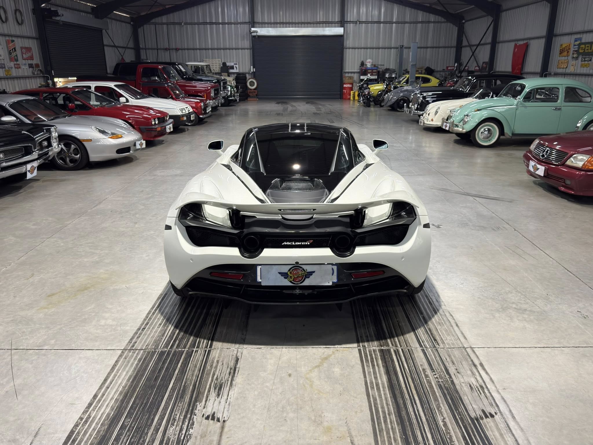 Rear view of a white McLaren sports car in a large showroom, flanked by rows of classic cars and vintage signs along the walls.