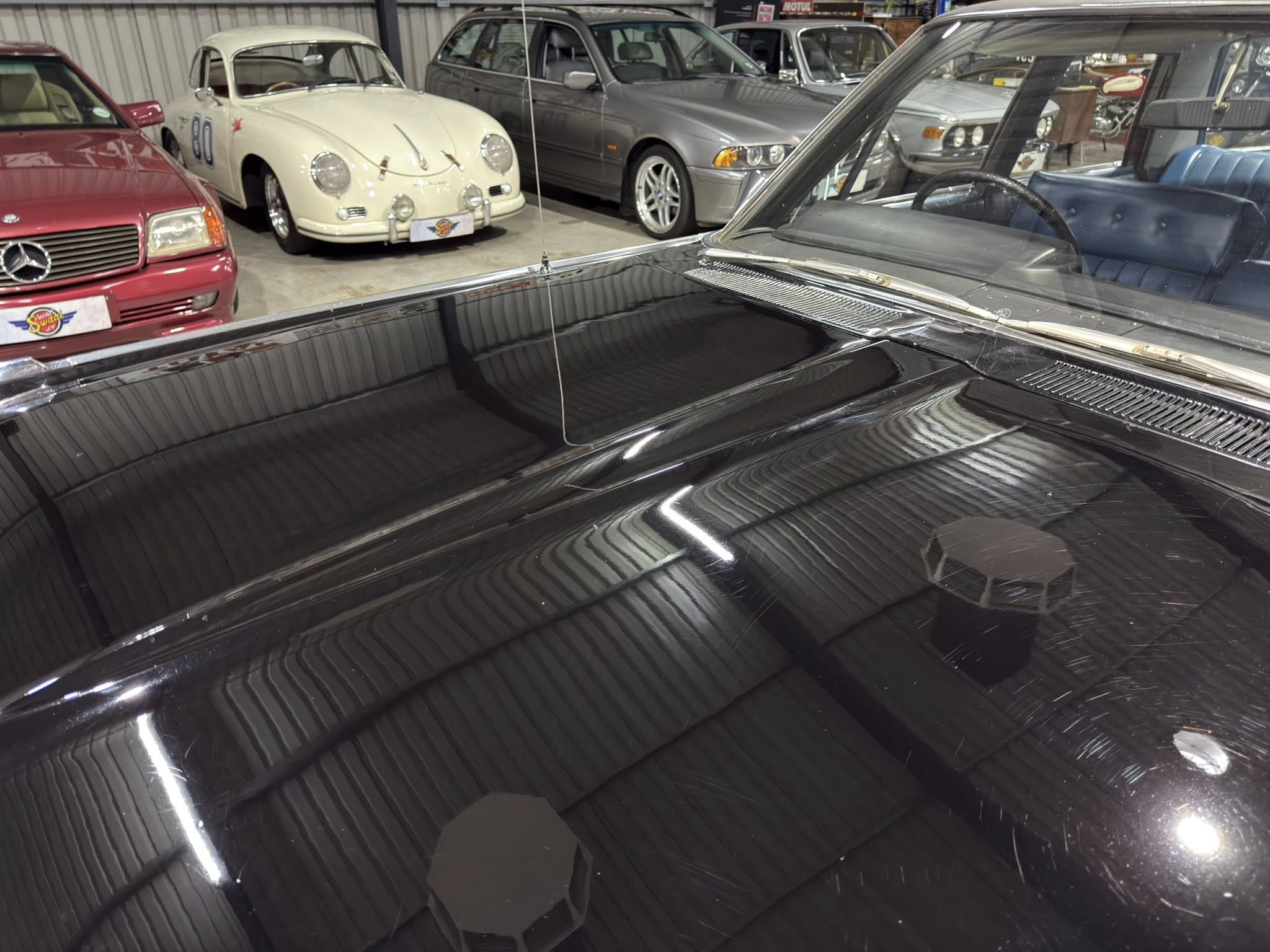 Interior shot of a classic car showroom with a glossy black hood in the foreground and vintage cars parked along the back wall, including a white relic and a silver sedan.