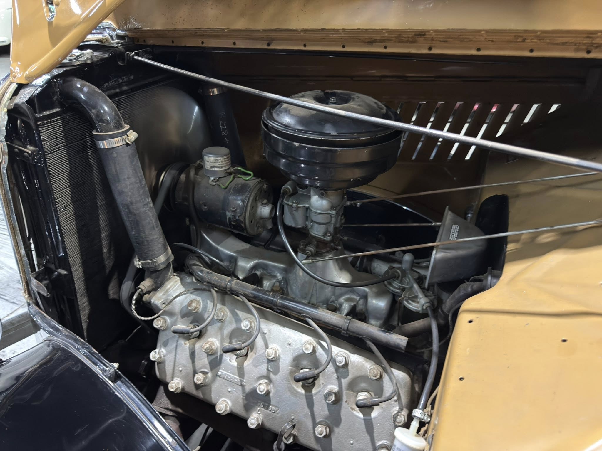 Open hood of a vintage car revealing its engine bay with a large grey engine block, hoses, and air filter housing; tan bodywork nearby.