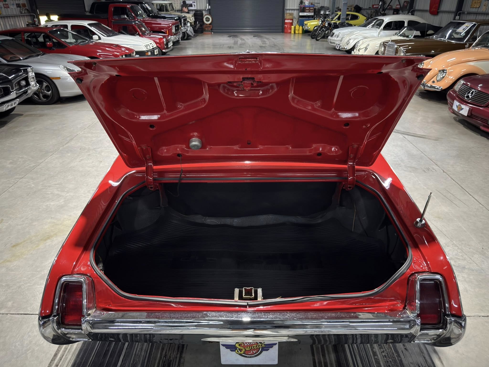 Open trunk of a red vintage car in a showroom filled with classic cars on both sides of the aisle.