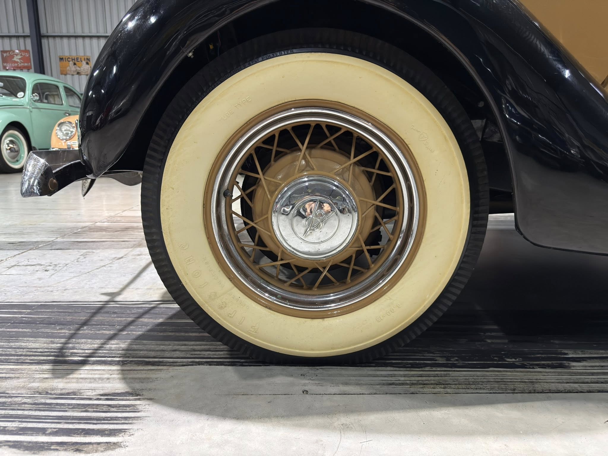 Close-up of a vintage car wheel with a beige wall tire and wire-spoke rim under a black fender in a showroom-like garage.