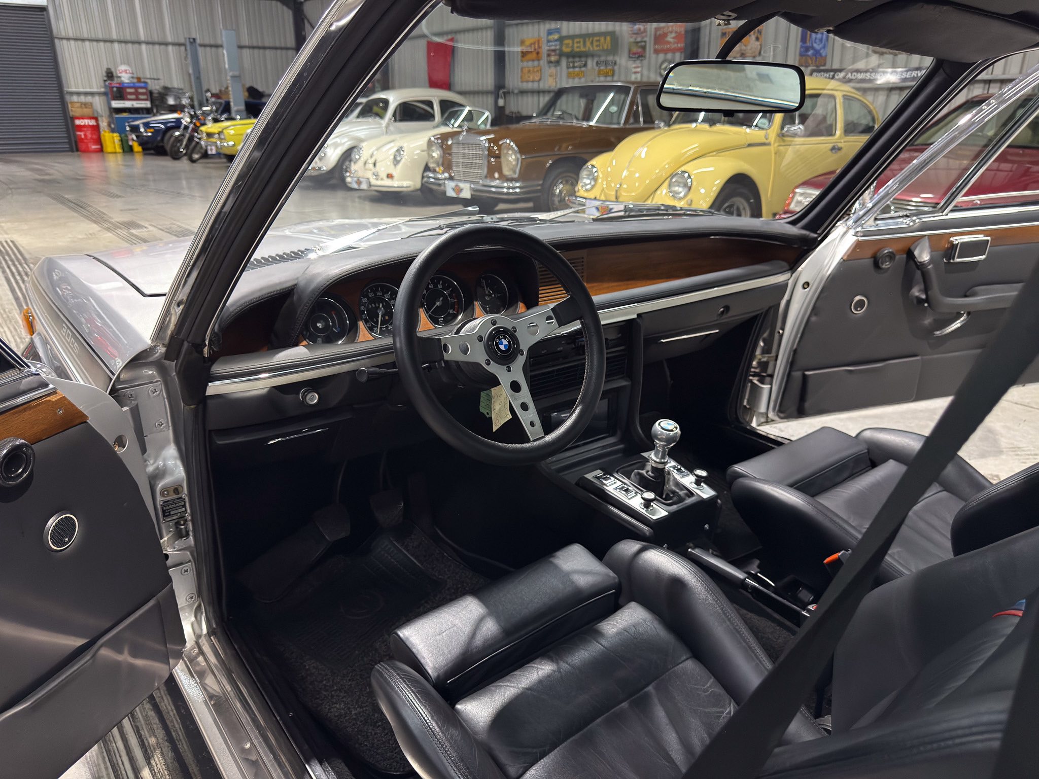 View from the driver’s seat of a classic car, showing black leather interior, wood trim, and a dashboard with analog gauges. Outside, vintage cars are visible in a showroom.