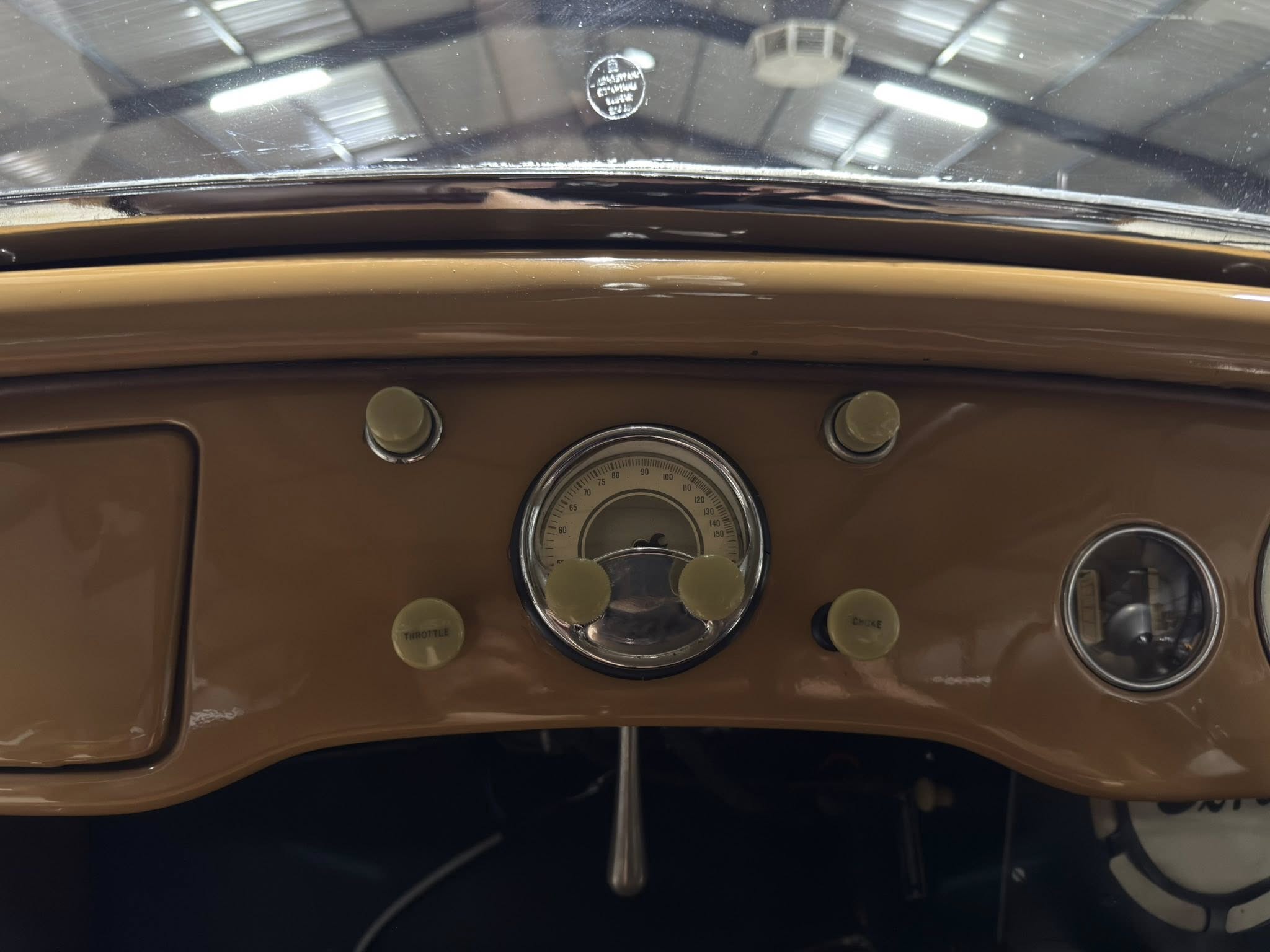 Close-up of a brown vintage car dashboard with a round gauge, beige knobs labeled throttle/check, and windshield reflections above.