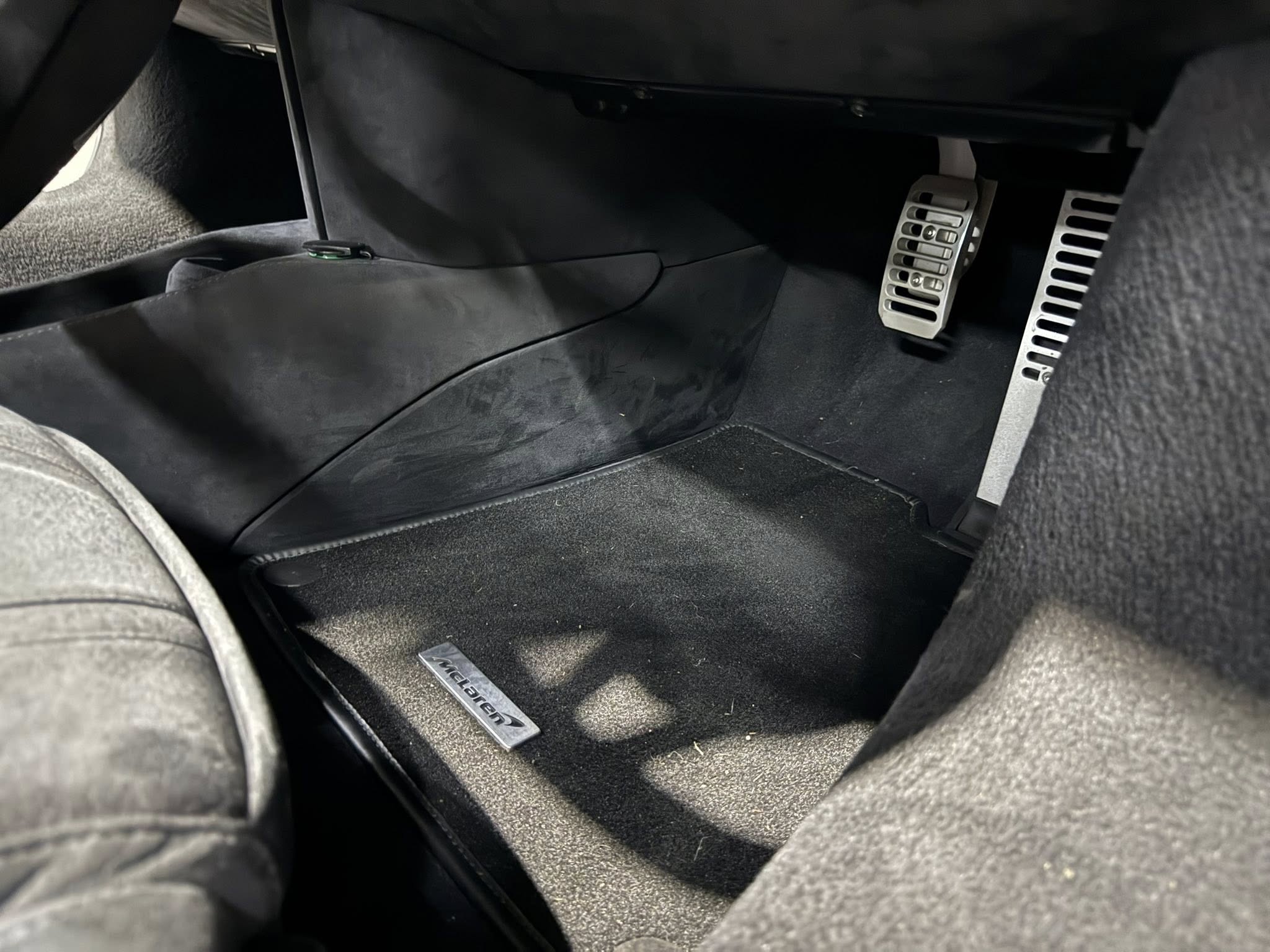 Interior of a dark luxury car showing the driver footwell with metal pedals and a branded McLaren floor mat seen on the carpeted floor.