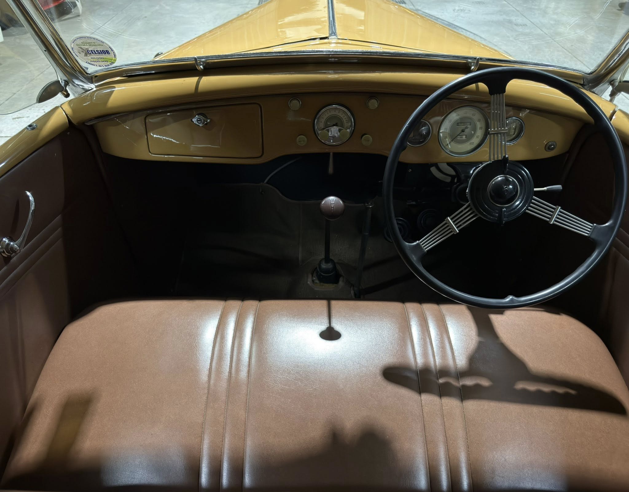 Vintage car interior with tan/brown leather seats and a large black steering wheel, chrome accents, and a tan dashboard viewed from the passenger seat.