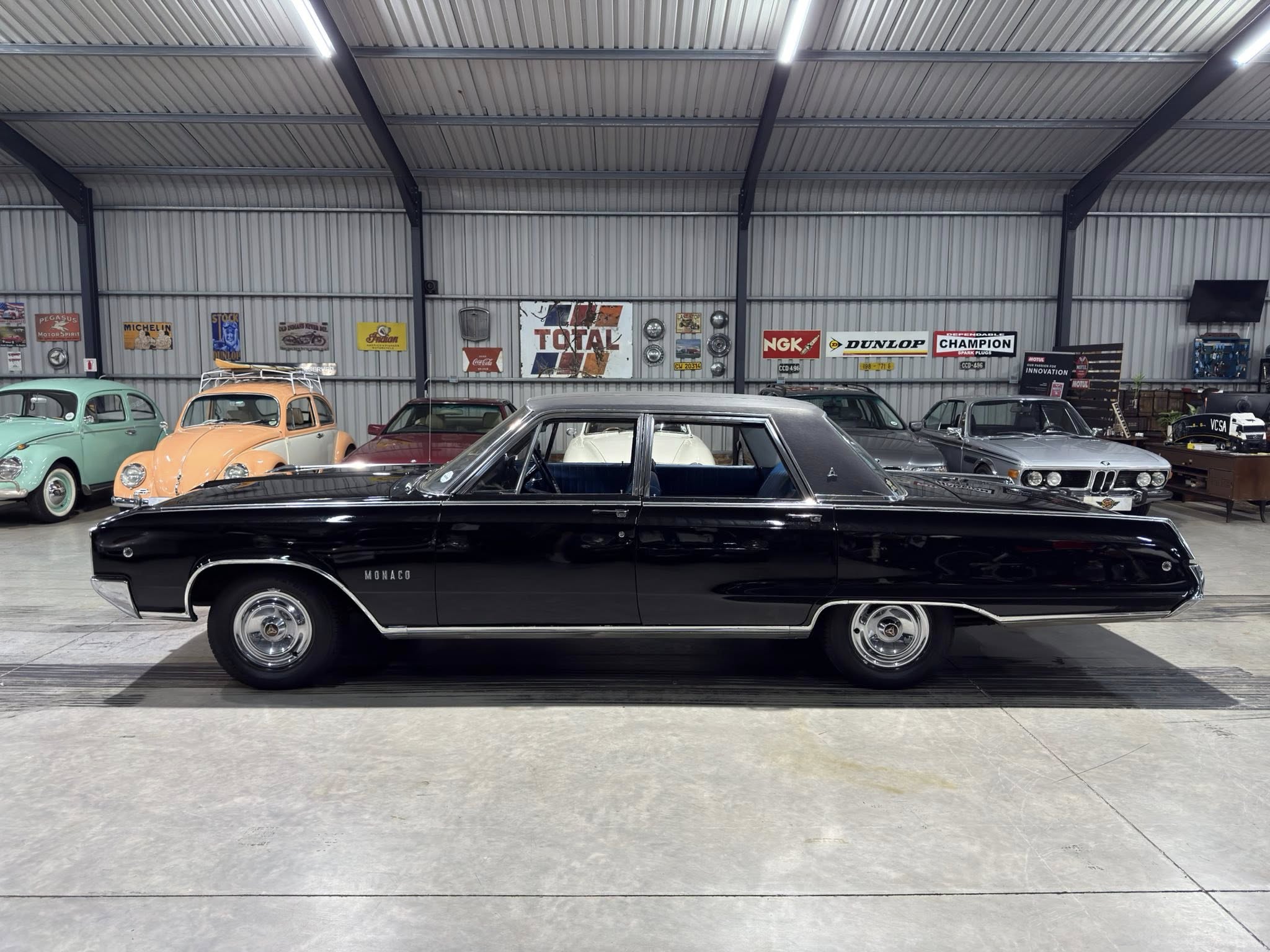 Long black vintage car in a showroom with pastel-colored cars and retro signs on a metal wall behind it, indoors titled Monaco on the side.