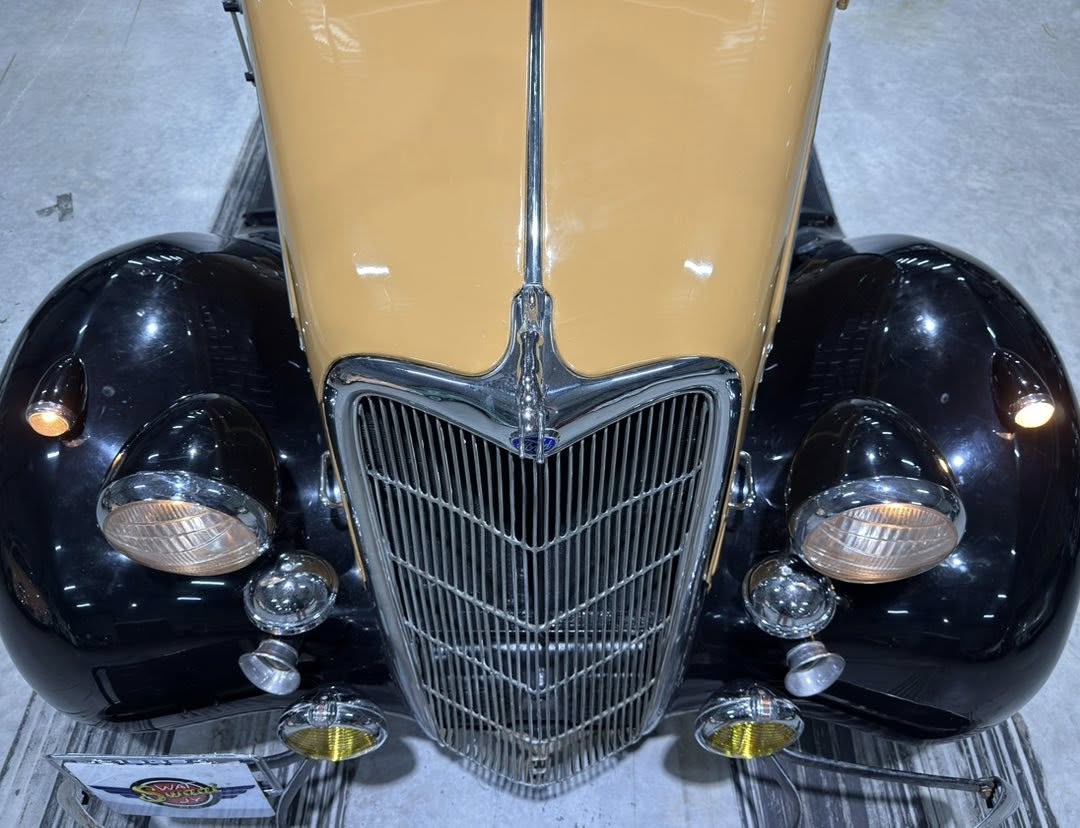 Front view of a vintage two-tone car (beige hood, black fenders) with a tall chrome grille and round headlights edge-to-edge on either side.