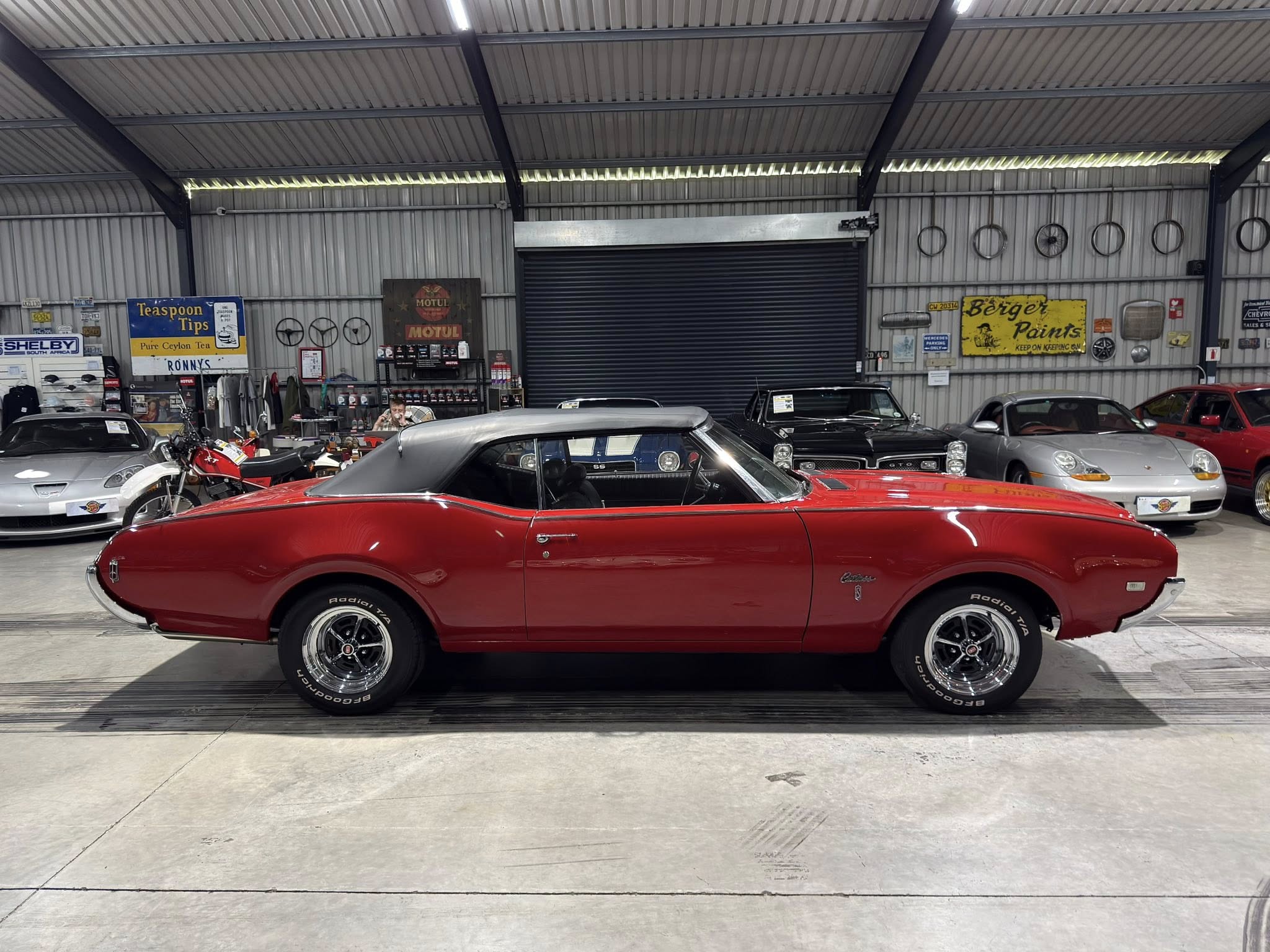 Red classic muscle car in a showroom foreground, with other vintage cars and retro signs on metal walls behind it.