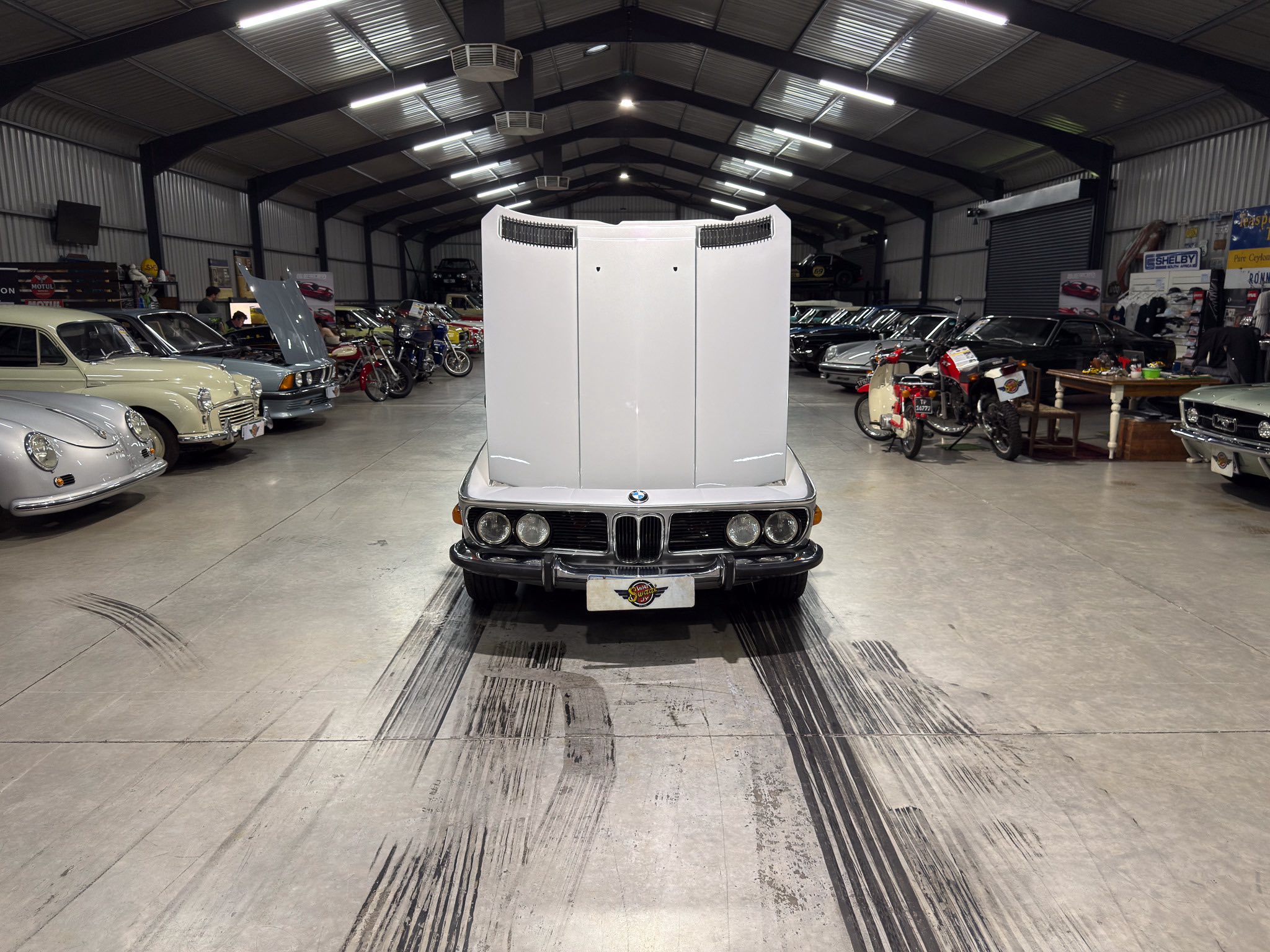 Classic car showroom with a white hood raised at center, surrounded by vintage cars and motorcycles.