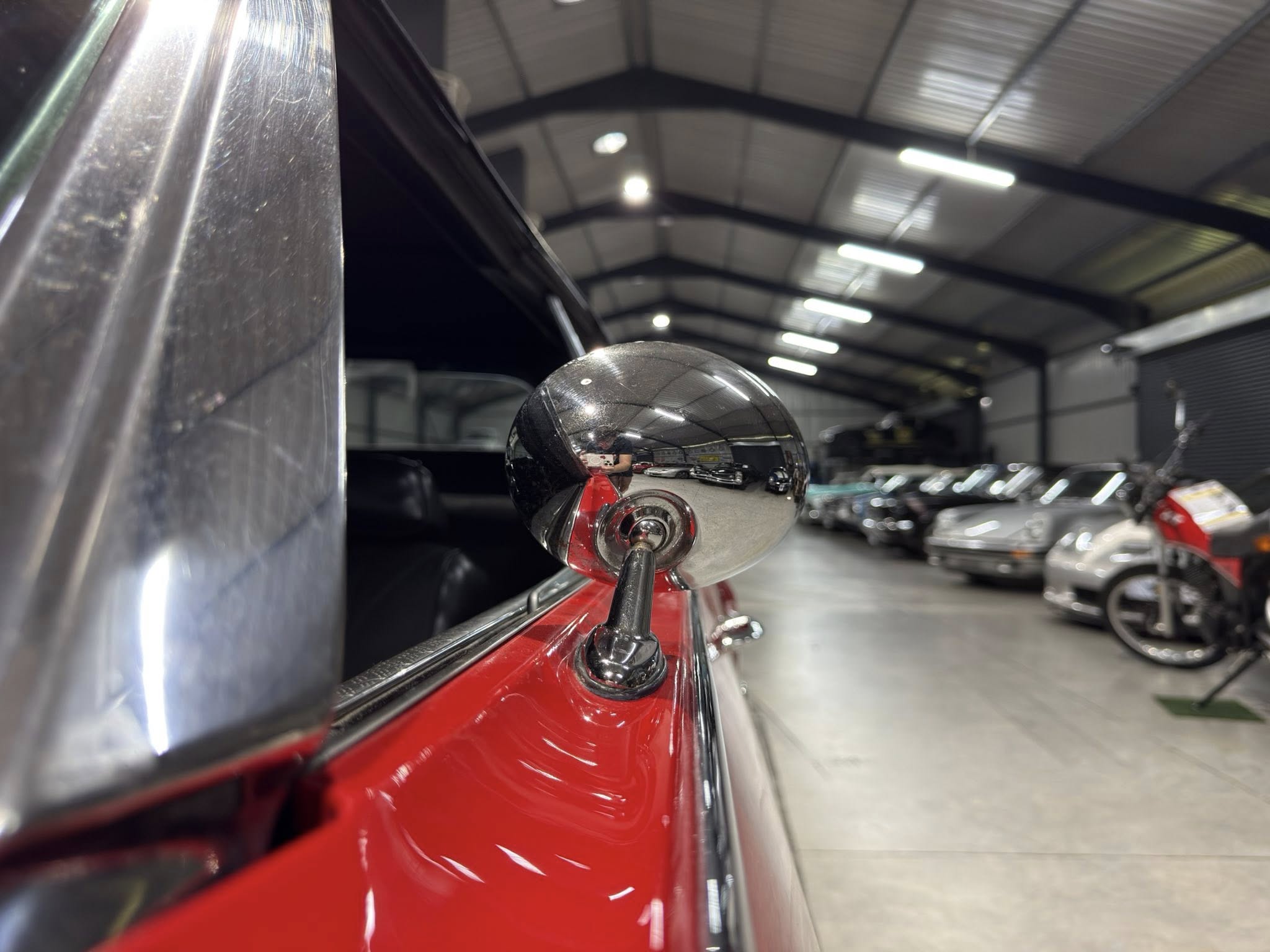 Close-up of a chrome side mirror on a red car inside a showroom with other vehicles in the background.