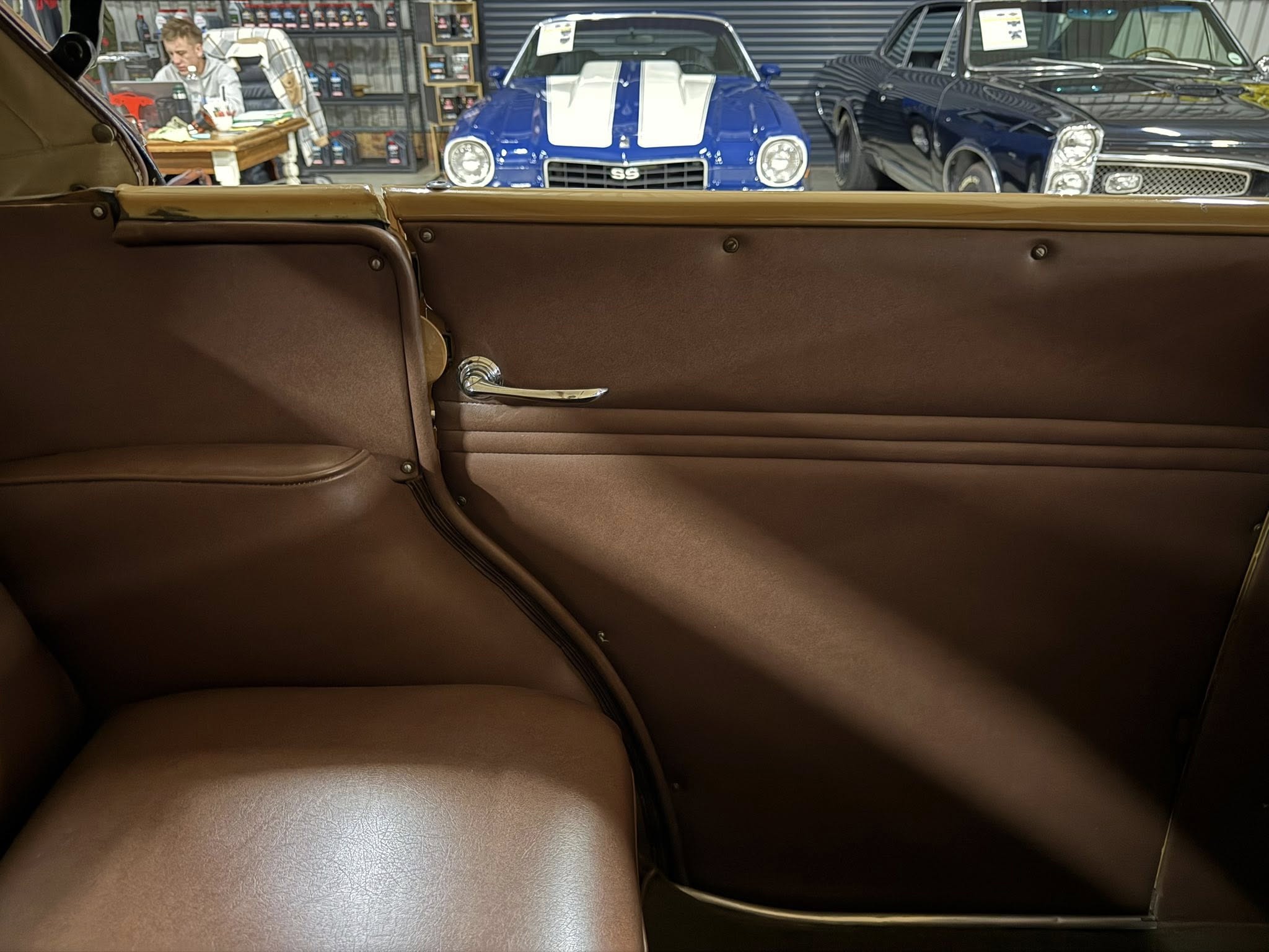 Interior view of a vintage brown leather car with a chrome door handle, looking out toward a blue classic car with white racing stripes in a showroom.