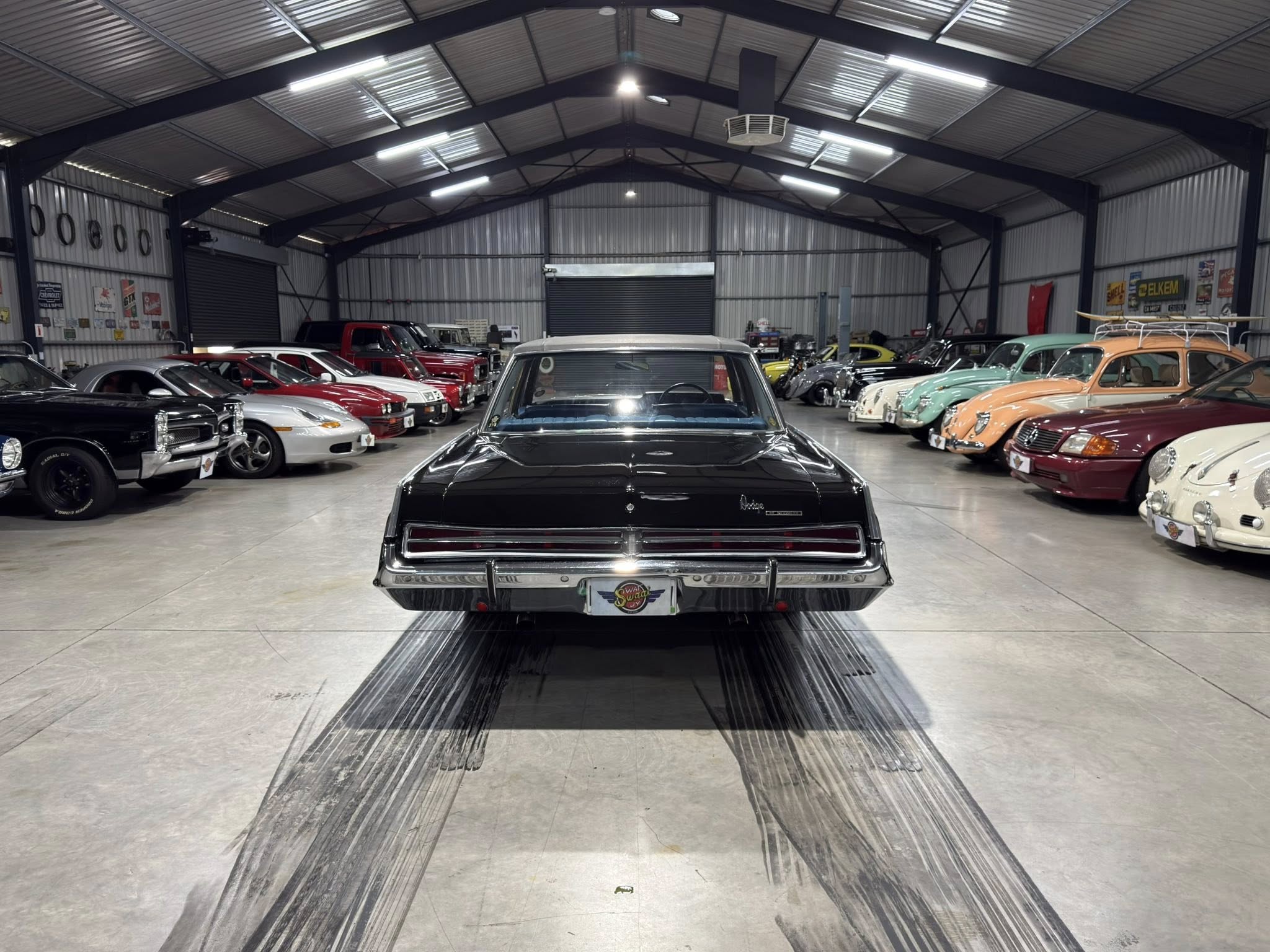Indoor classic car showroom with a line of vintage cars on both sides and a dark blue ceiling girders.