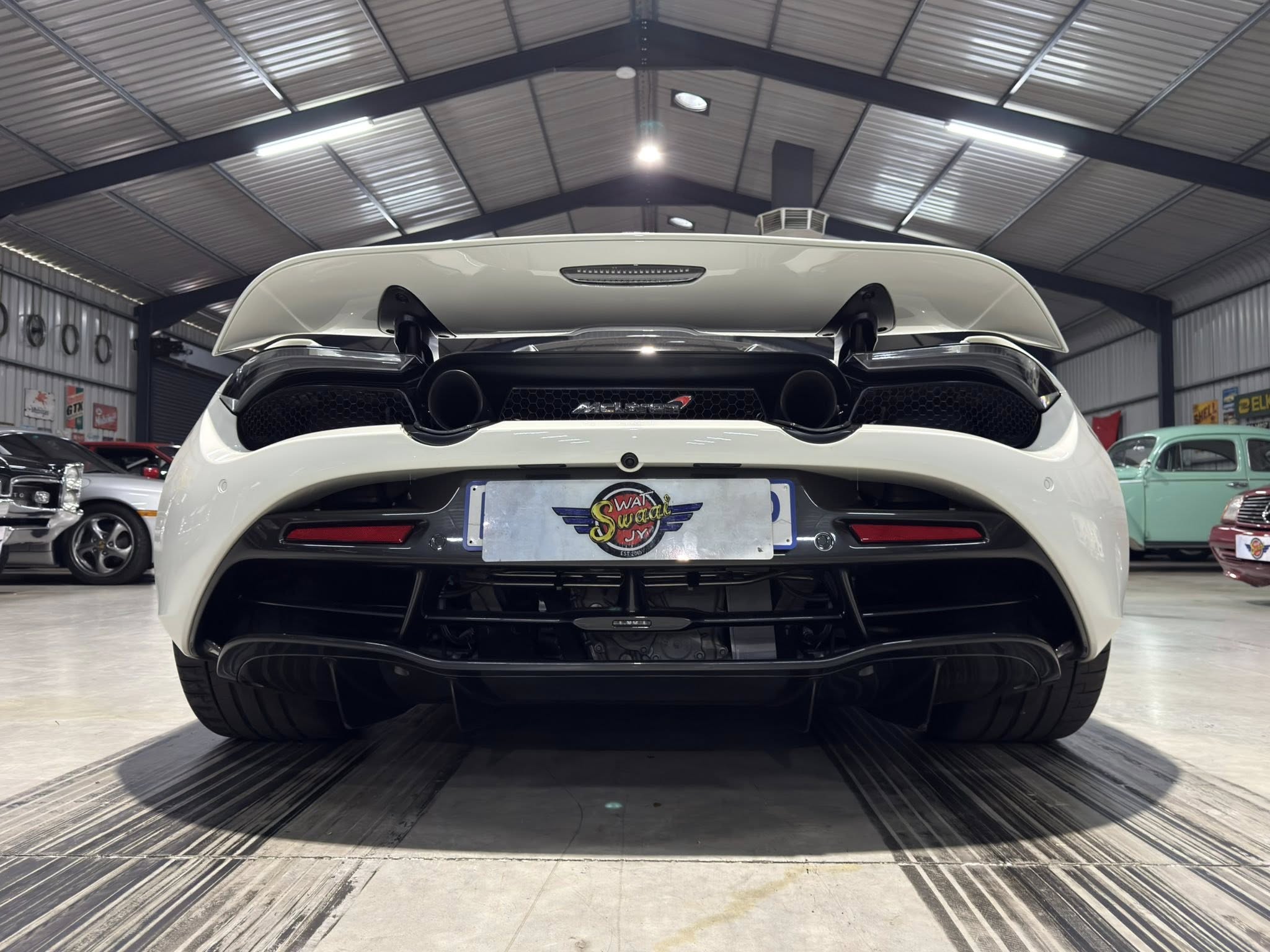Rear view of a white sports car inside a showroom, large rear spoiler and dual exhausts visible before a license plate area.