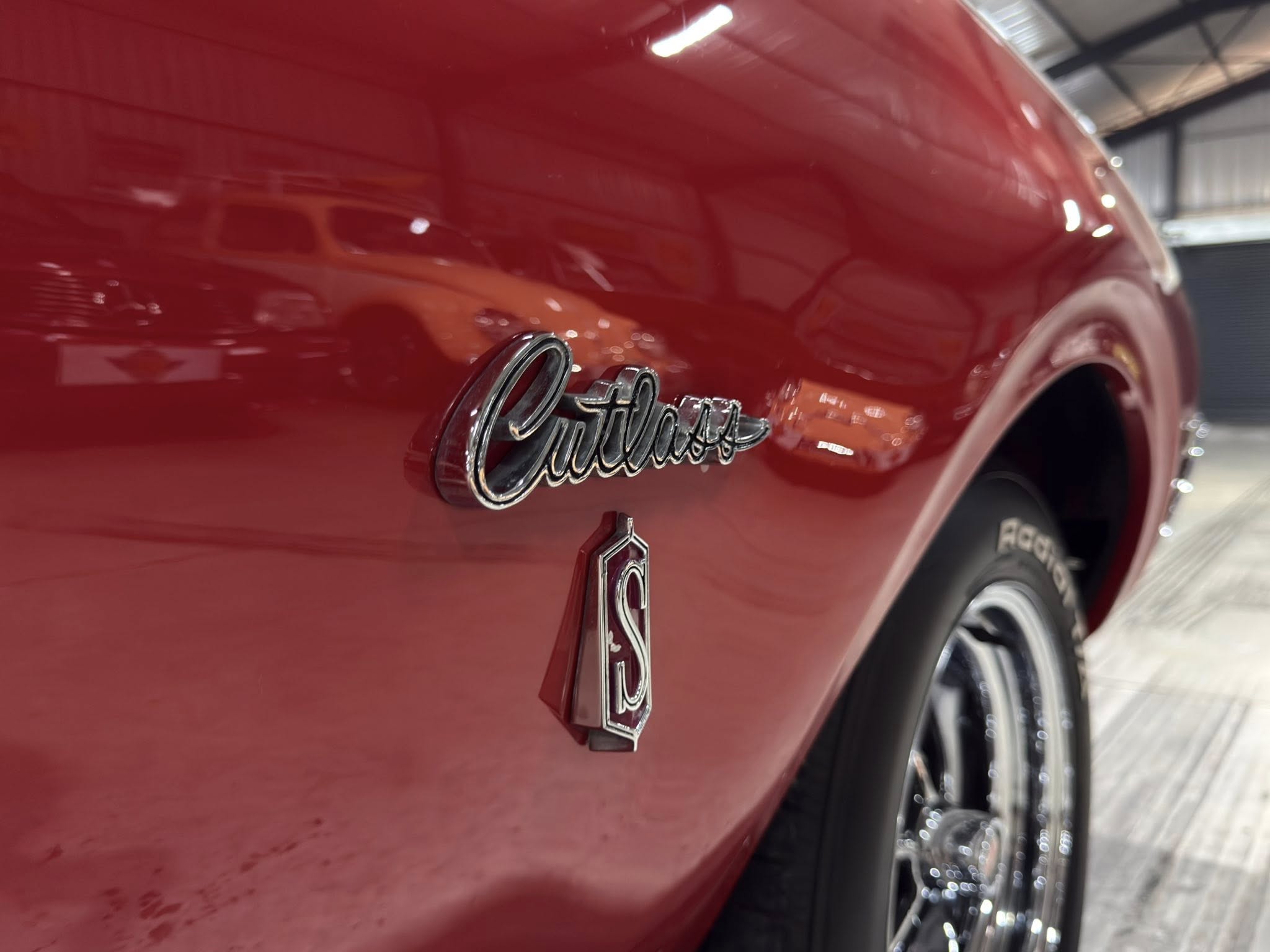 Close-up of a red car body with a chrome, cursive badge and a wheel visible in the background.