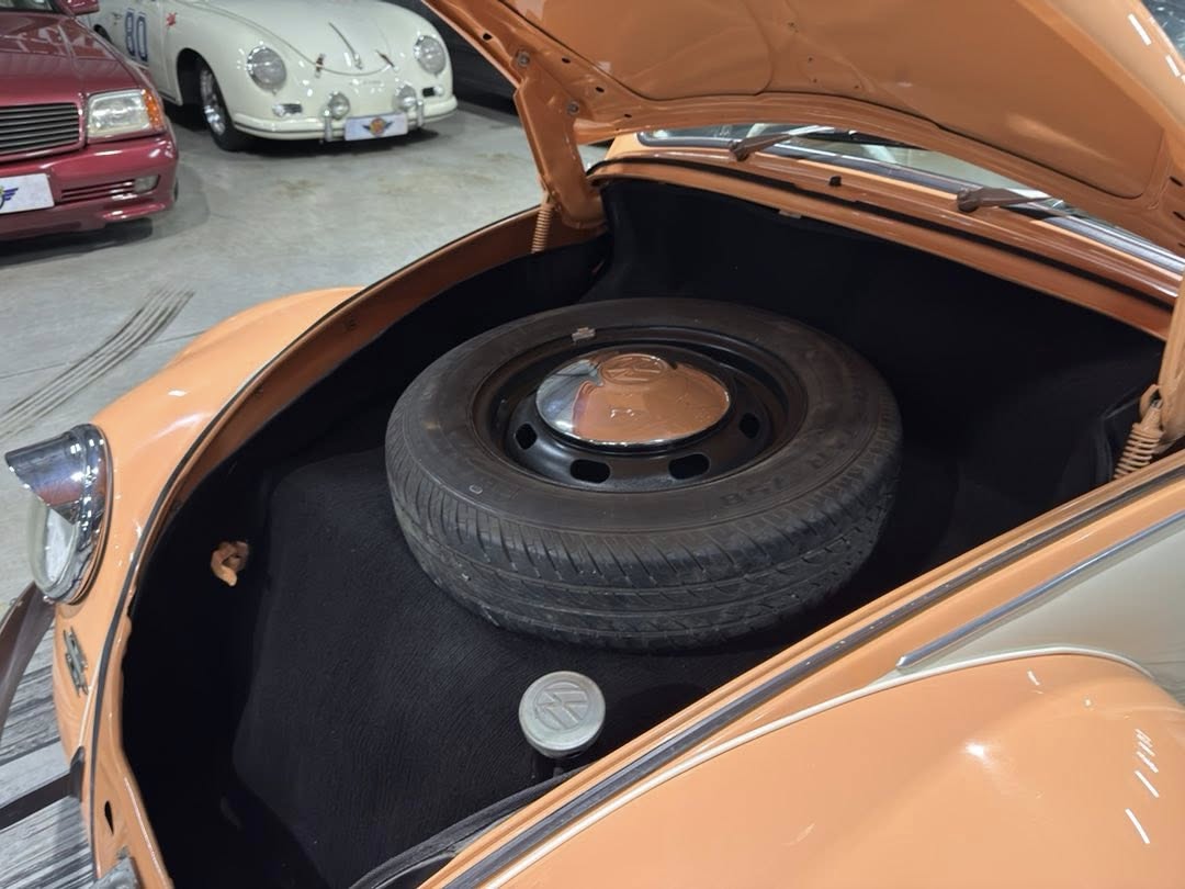 Orange classic car with its trunk open revealing a mounted spare tire in the storage area, parked in a showroom with other vintage cars in the background.