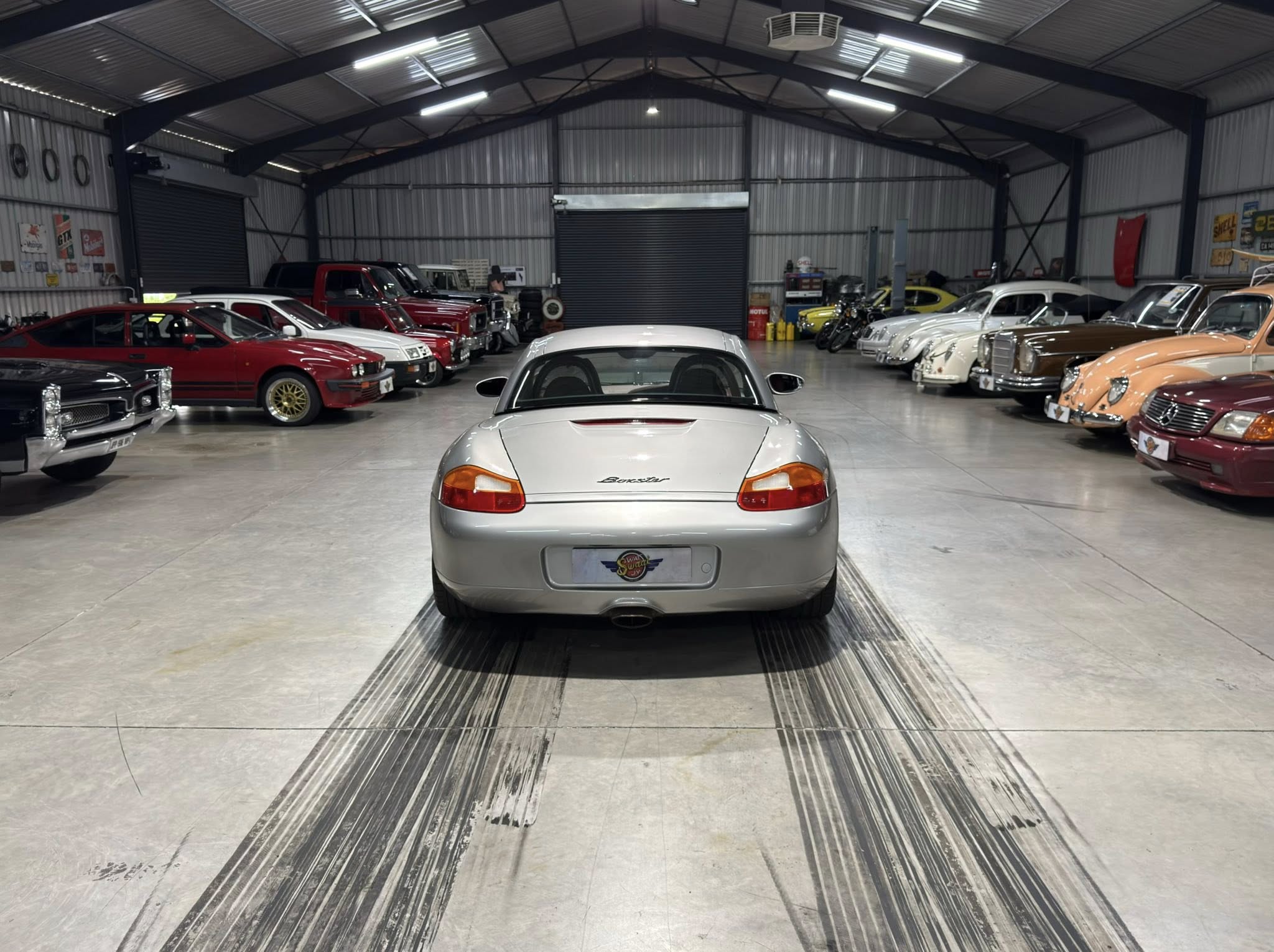 Rear view of a silver Porsche Boxster in a car showroom, with a row of classic cars along both sides in a well-lit warehouse.