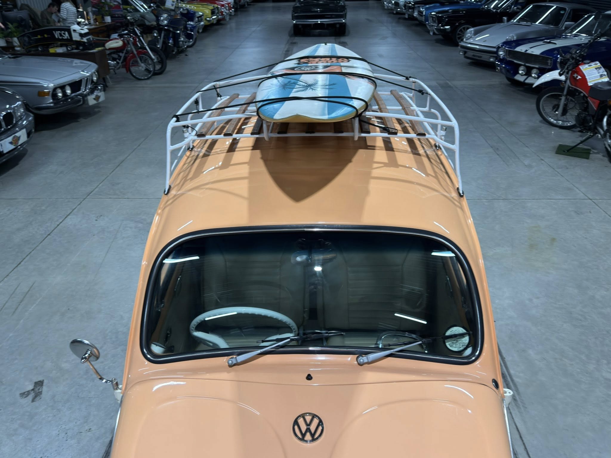 Top-down view of an orange vintage VW in a showroom, with a blue-and-white surfboard on its roof rack, flanked by rows of classic cars and motorcycles.