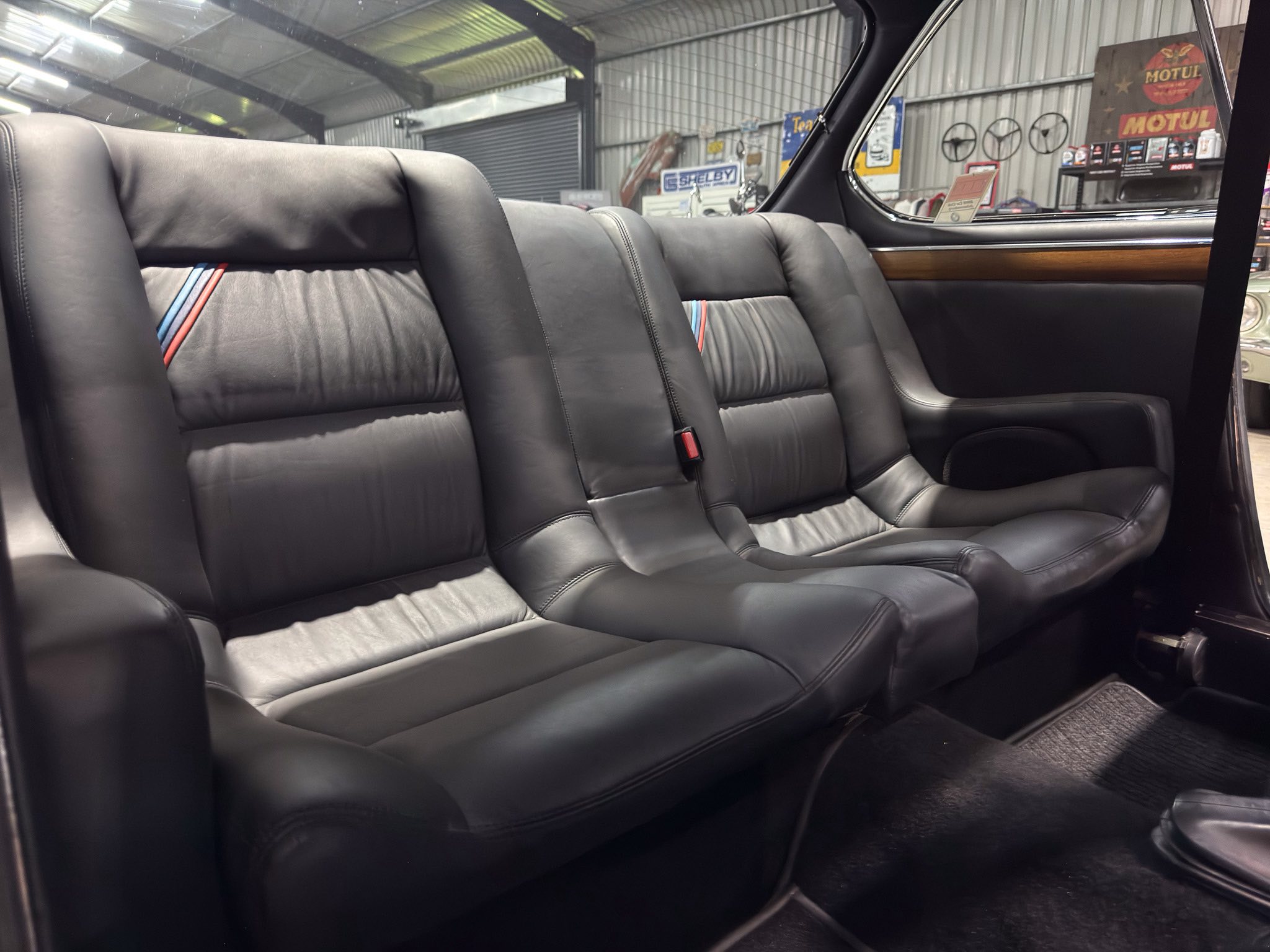 Rear row of black leather car seats with wood trim, seen from the back seat, inside a showroom or garage. Outside view shows workshop signs.