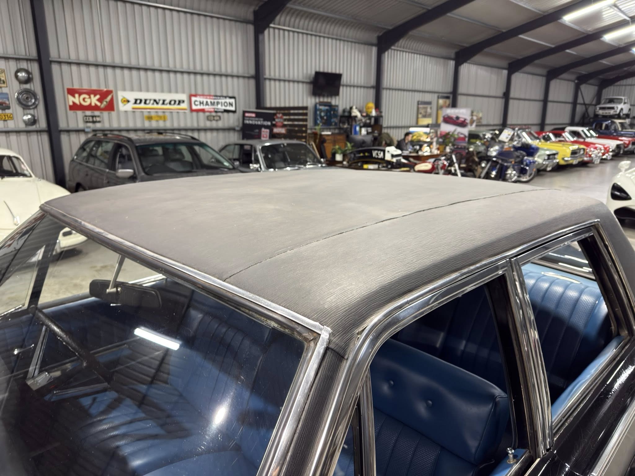 Indoor garage displaying vintage cars, metal walls, and sponsor banners (NGK, Dunlop, Champion) on the back wall.