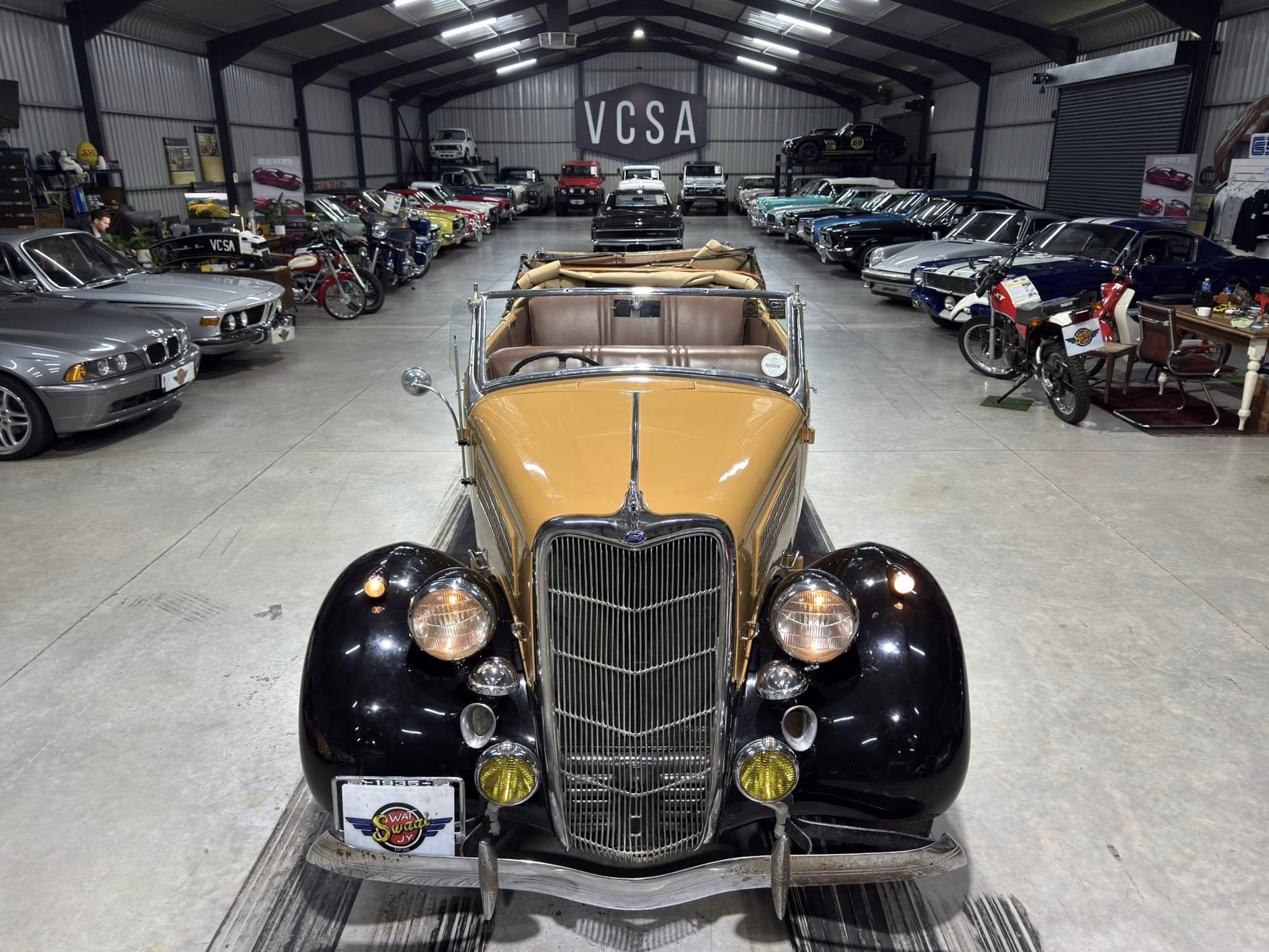 Front view of a tan vintage convertible inside a car showroom, with rows of classic cars on both sides and a VCSA sign overhead, creating a museum-like atmosphere.