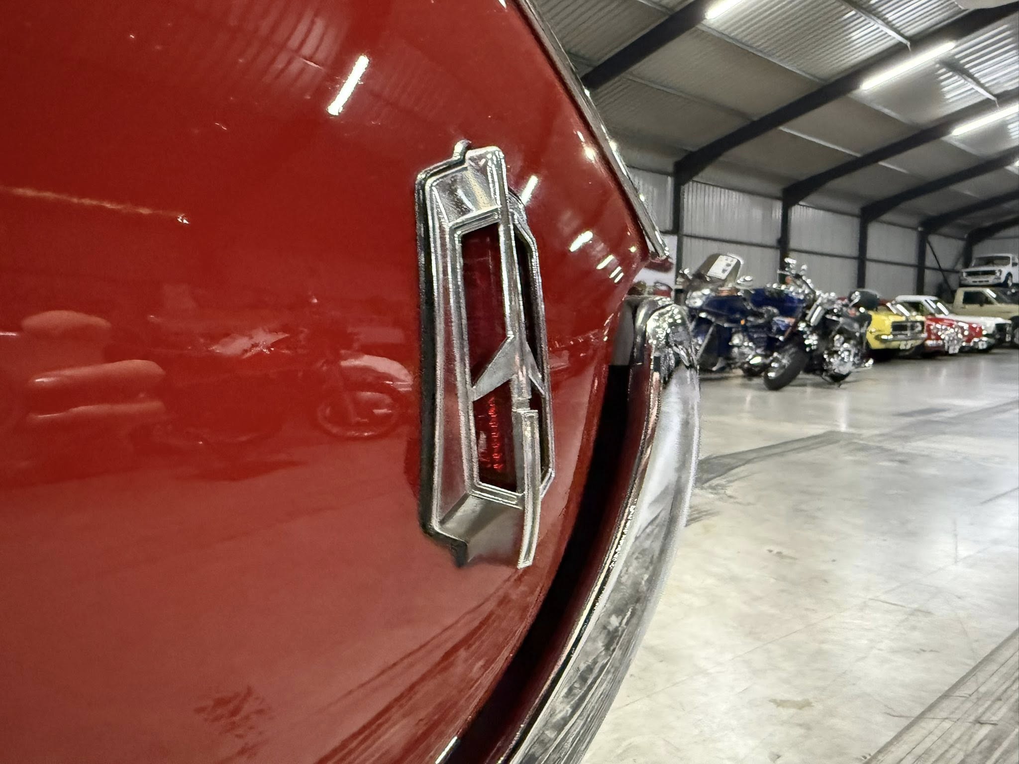 Close-up of shiny chrome trim on a red classic car, with a showroom containing motorcycles in the background.