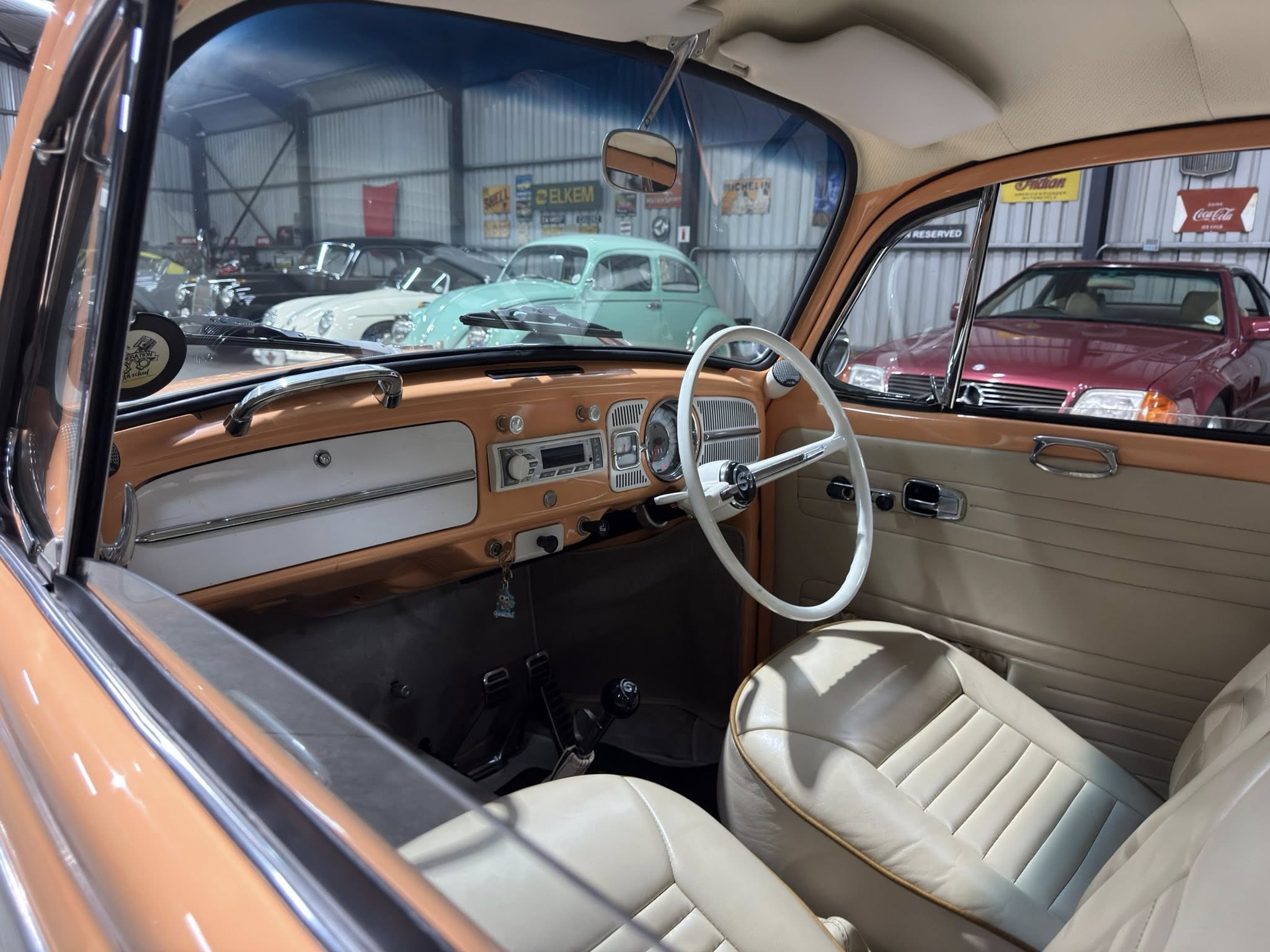 Interior of a vintage orange car with cream seats and white steering wheel, showroom of classic cars visible through the windshield.
