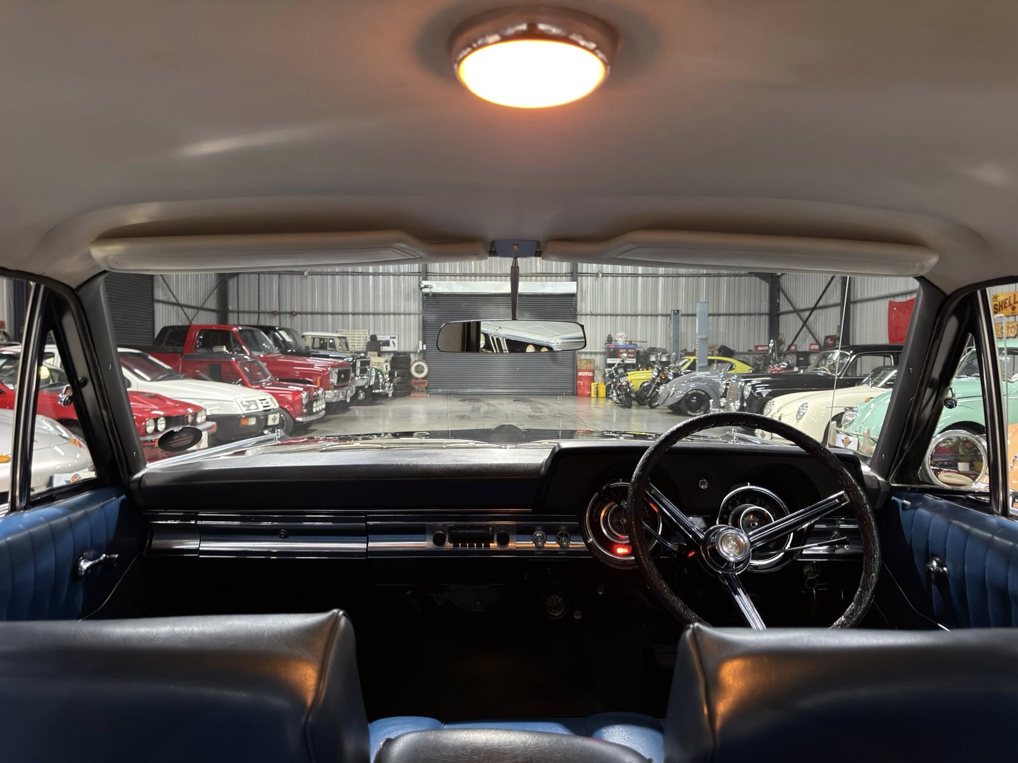 View from the driver’s seat of a vintage car, with the steering wheel and dash visible, showing a showroom full of classic cars in the background.