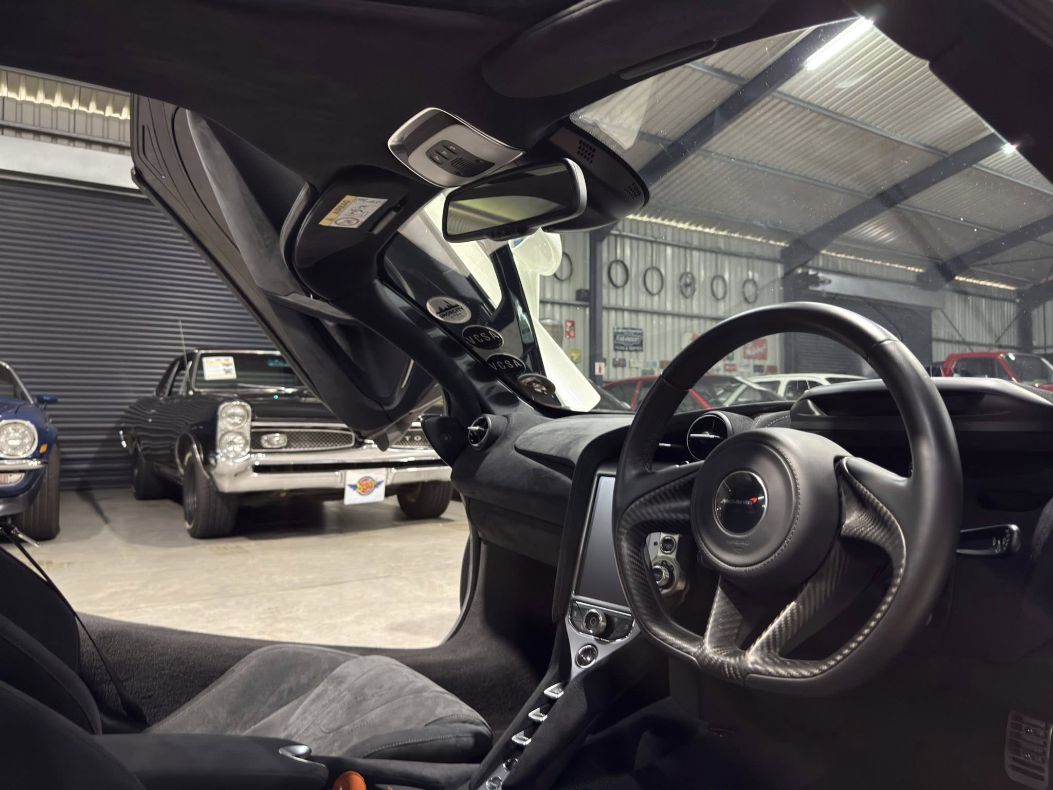 Right-hand-drive car interior with steering wheel, carbon-fiber dash, and rear-view mirror; showroom with classic cars outside the windshield.