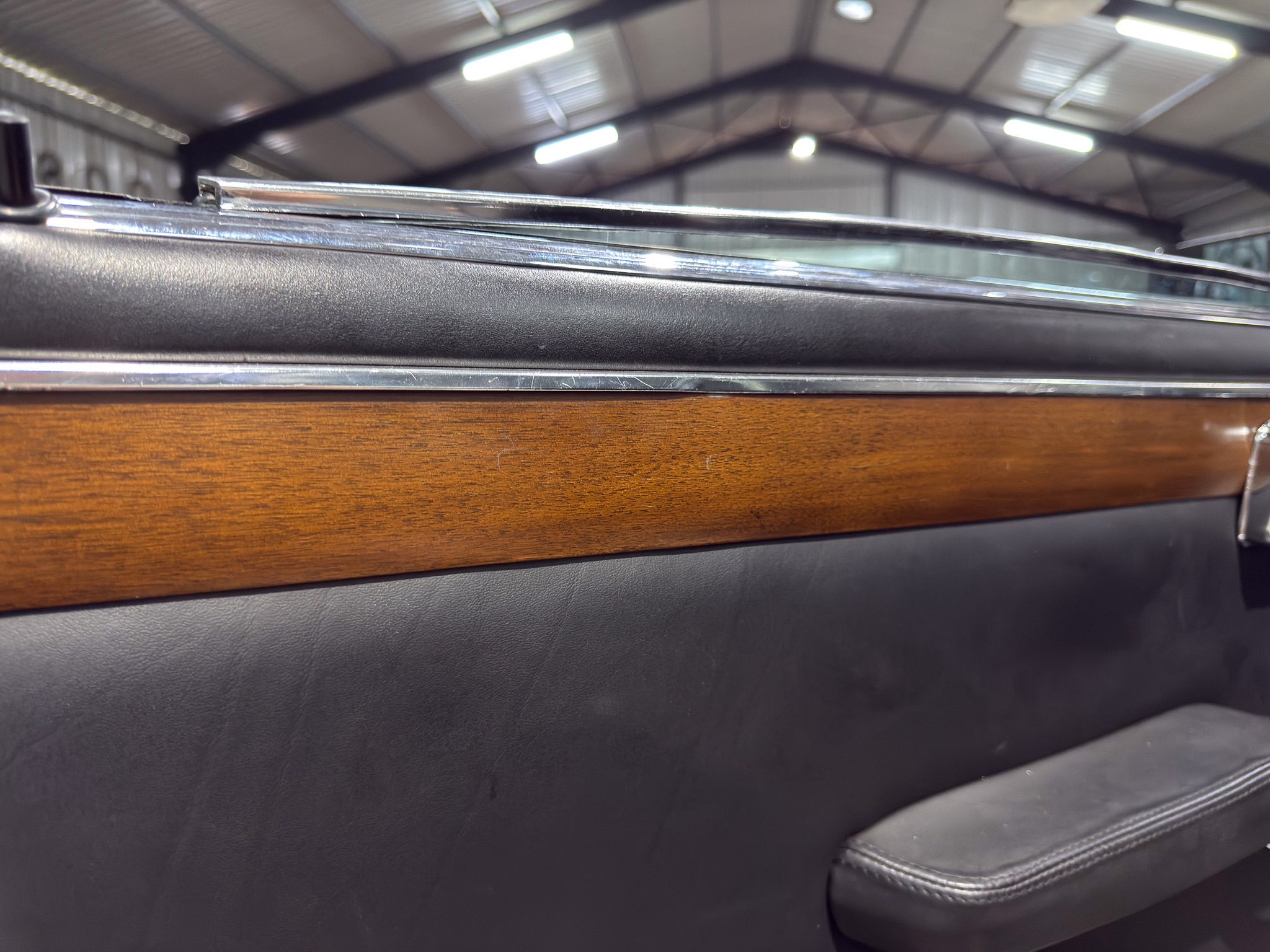 Close-up of a car door interior showing black padded armrest, chrome trim, and a wood panel with a garage-like background.