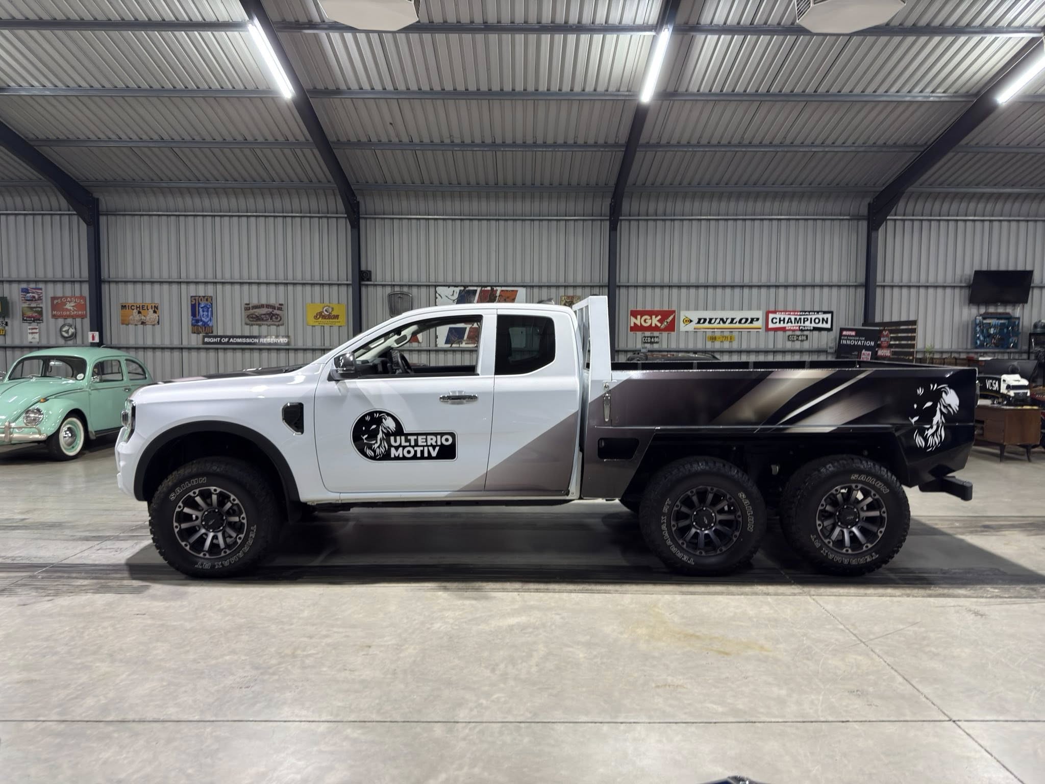 White pickup truck in a showroom with a vintage green car on the left and retro signs on the wall behind it